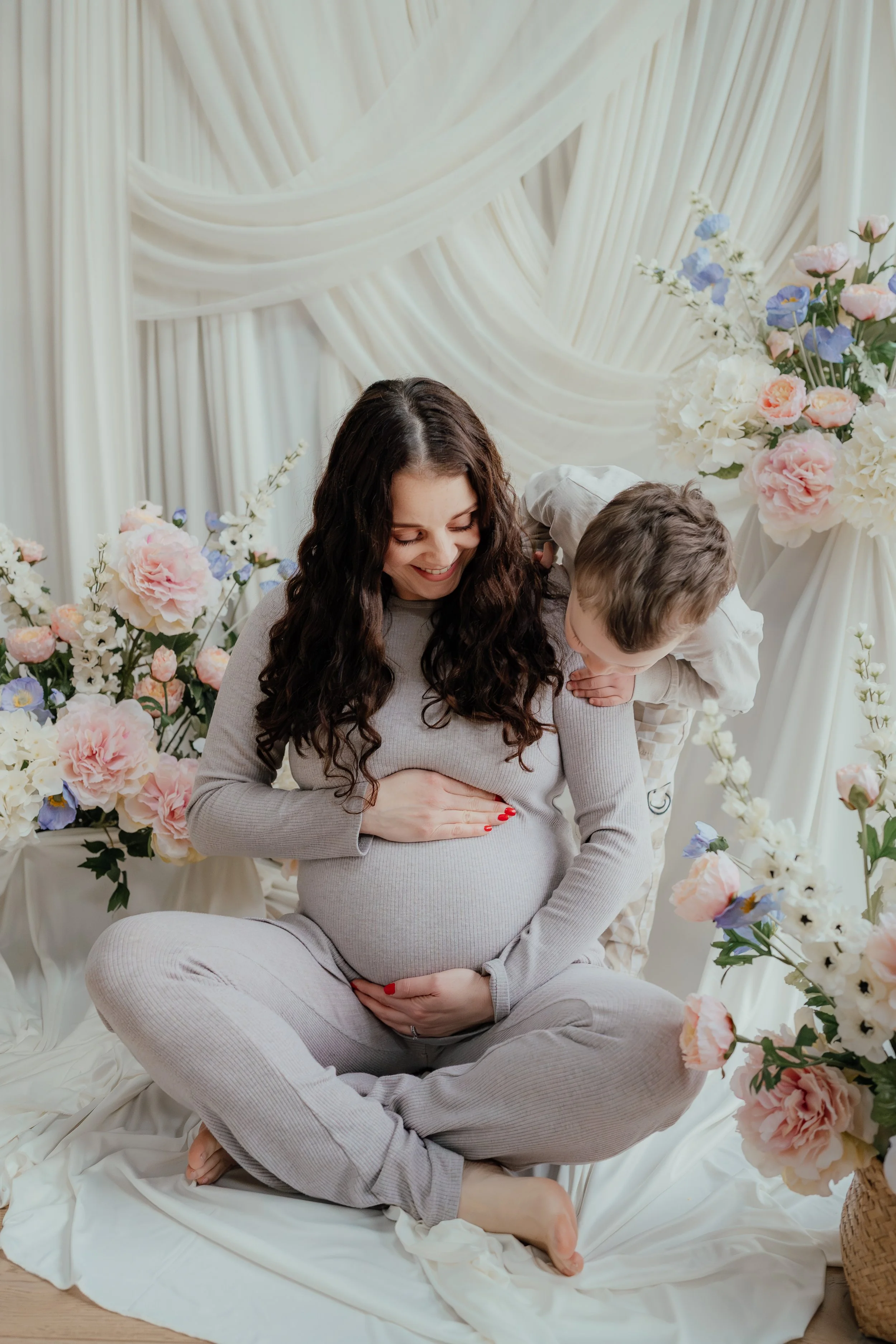 pregnant mum with long dark hair with her son against a white draped backdrop with spring flowers