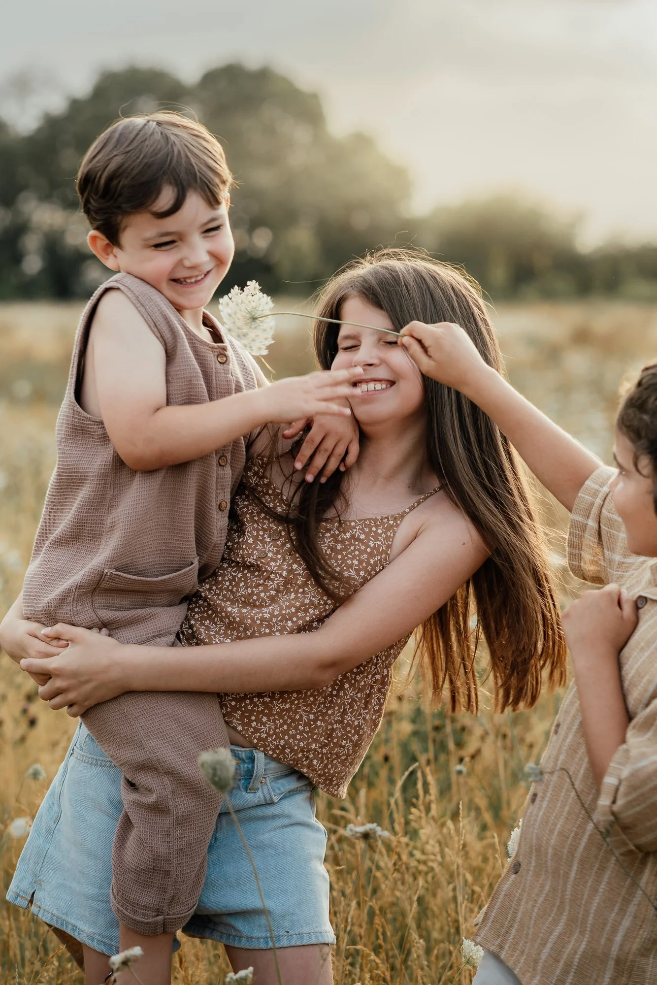 three siblings in summer clothing are laughing in a meadow at sunset with one boy tickling the other with a dandelion, whilst being held in his big sister's arms