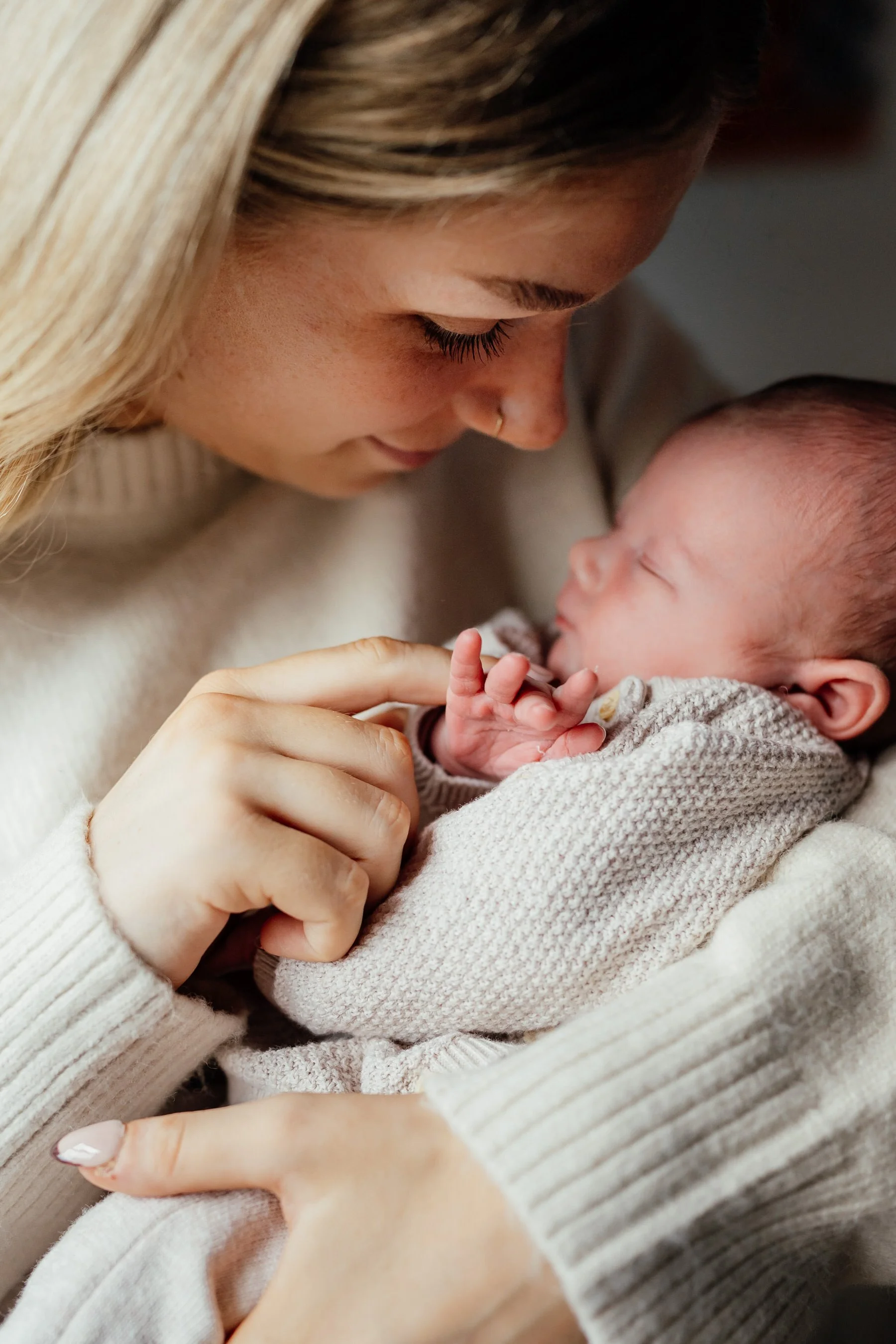 mum with blonde hair wearing a cream jumper holds her newborn baby girl in her arms, touching the baby's tiny hand with her finger