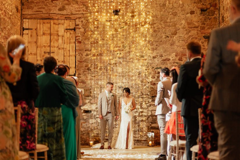 bride and groom holding hands, about to walk back down the aisle after getting married, with a fairy light backdrop in a barn