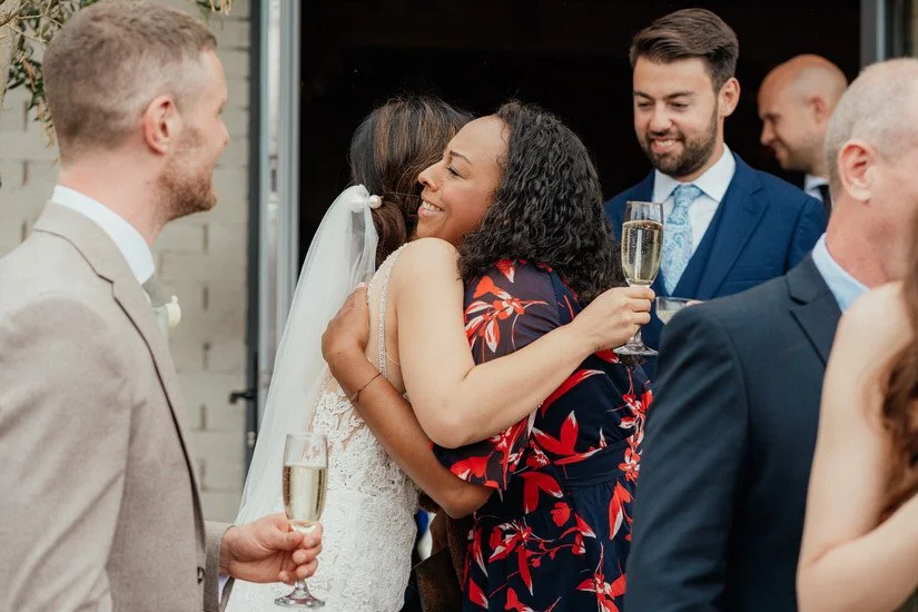 bride hugging a friend in a red and dark blue floral dress at a wedding drinks reception