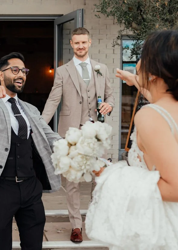 bride about to hug her friend in a grey suit, with the groom in the background smiling