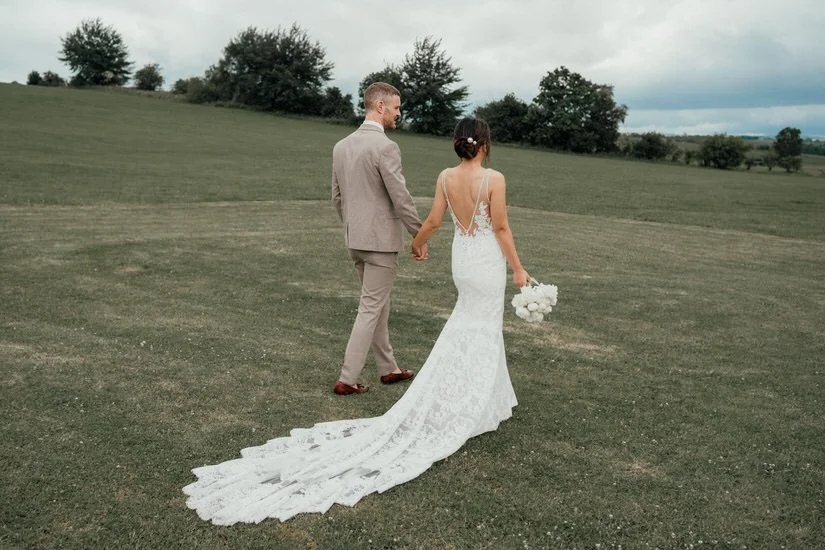 bride &amp; groom holding hands, walking away through a green field
