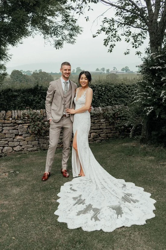 bride in a lace dress with long train and groom in a garden with a stone wall behind