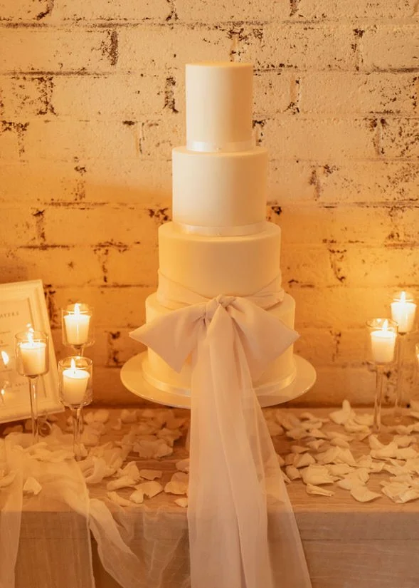 elegant minimal white 4 tier wedding cake with a white ribbon around the bottom tier, lit with candles either side