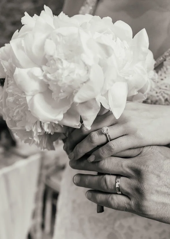 black and white photo of a peony bridal bouquet with the bride &amp; grooms wedding rings on their hands