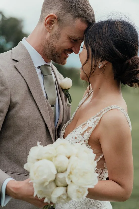 bride &amp; groom smiling, with their foreheads touching, with a bouquet of white peonies in the foreground