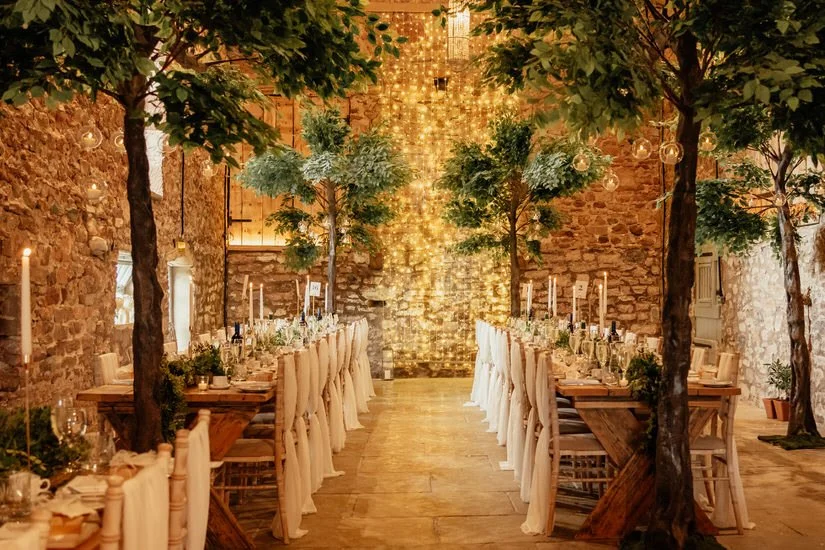 Eden Barn wedding venue dressed for a wedding breakfast with long trestle tables, fairy light backdrop and indoor green trees