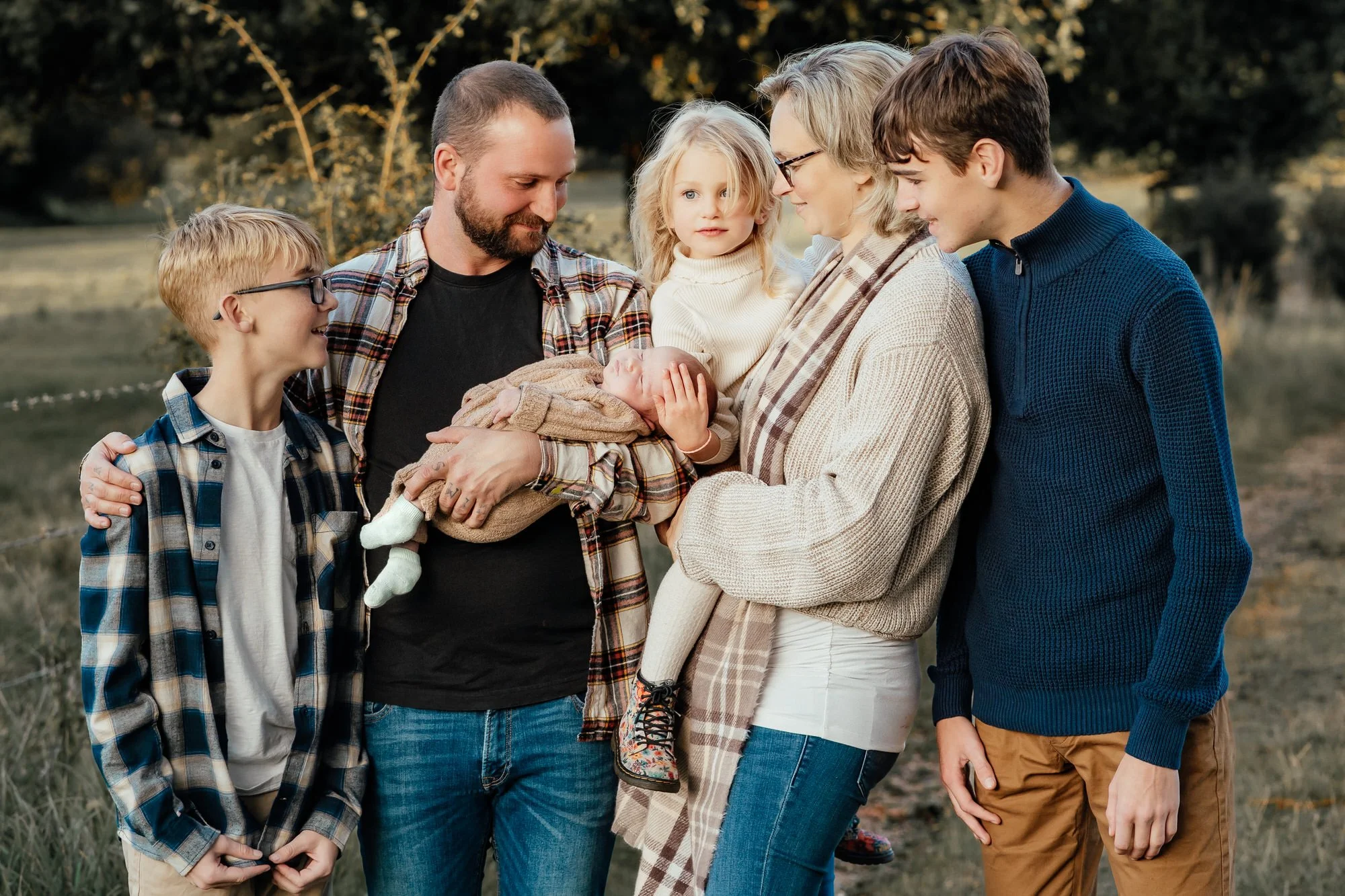 larger family with 2 older teenage boys, 1 young girl and 1 newborn baby being cuddled with mum and dad smile at the baby with the park in the background