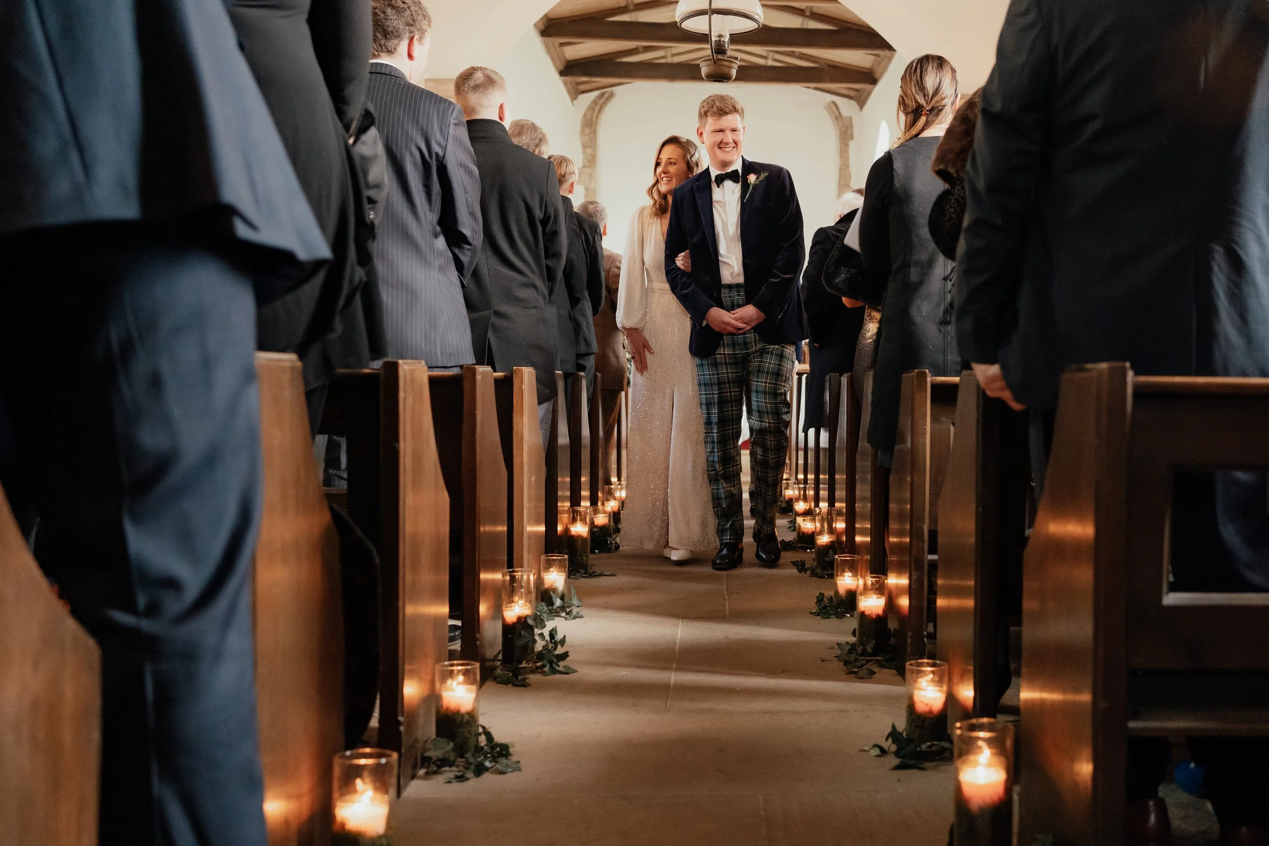 bride & groom in tartan trousers walking back down chapel aisle lit by candles