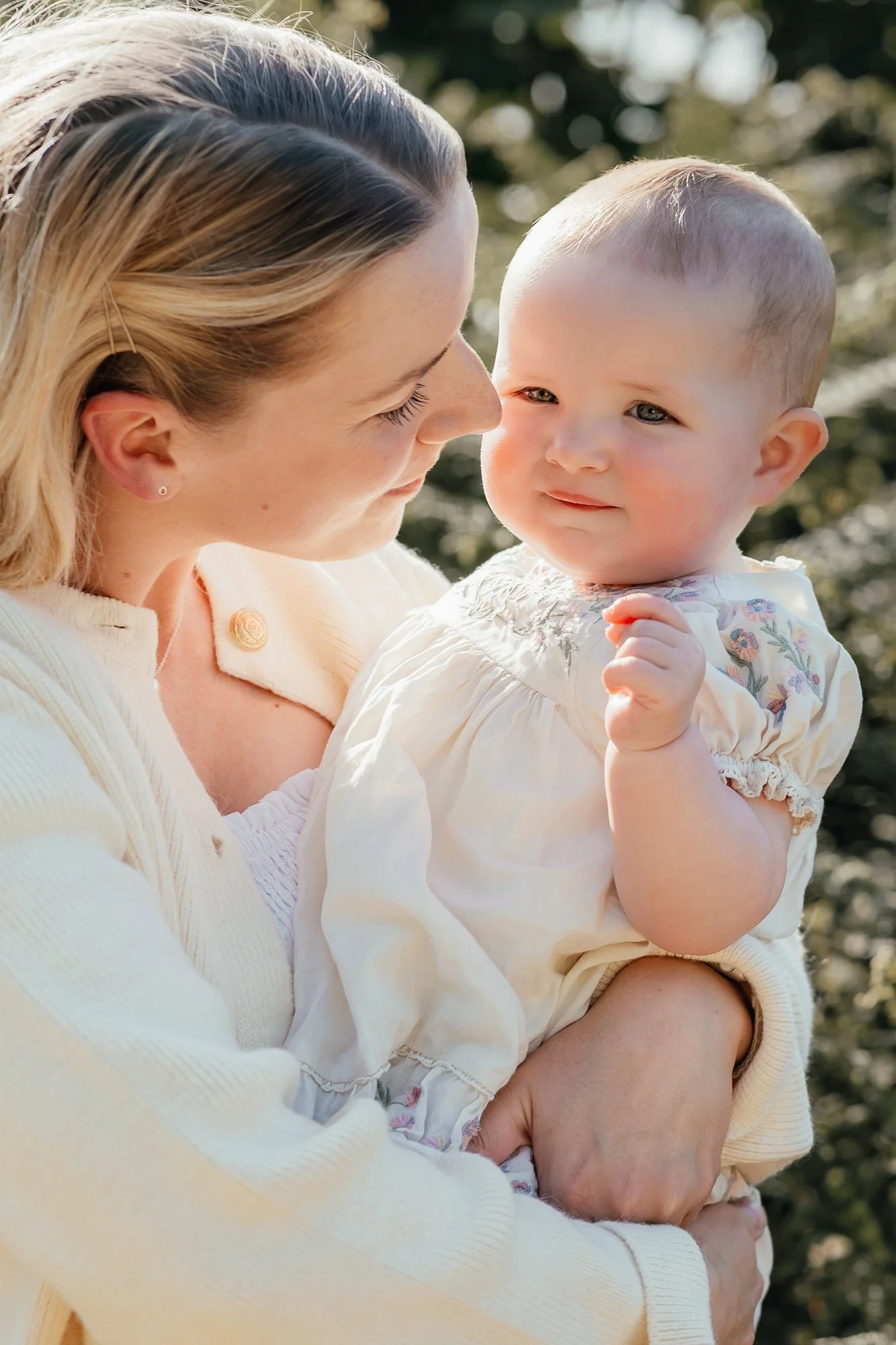 mother wearing a cream cardigan looks fondly at her 1yr old baby girl in a summer floral dress outside in the summer