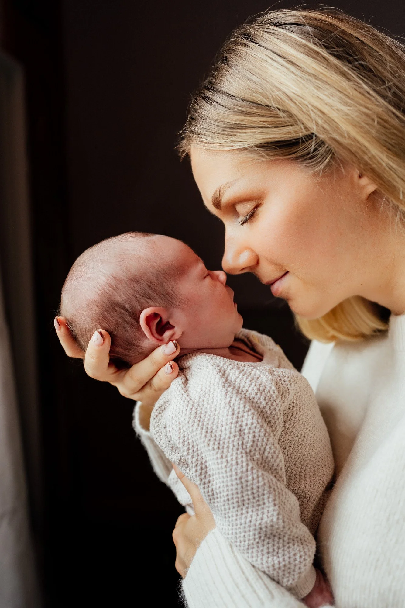mother holds her newborn baby in her hands with noses touching and eyes closed against a dark background