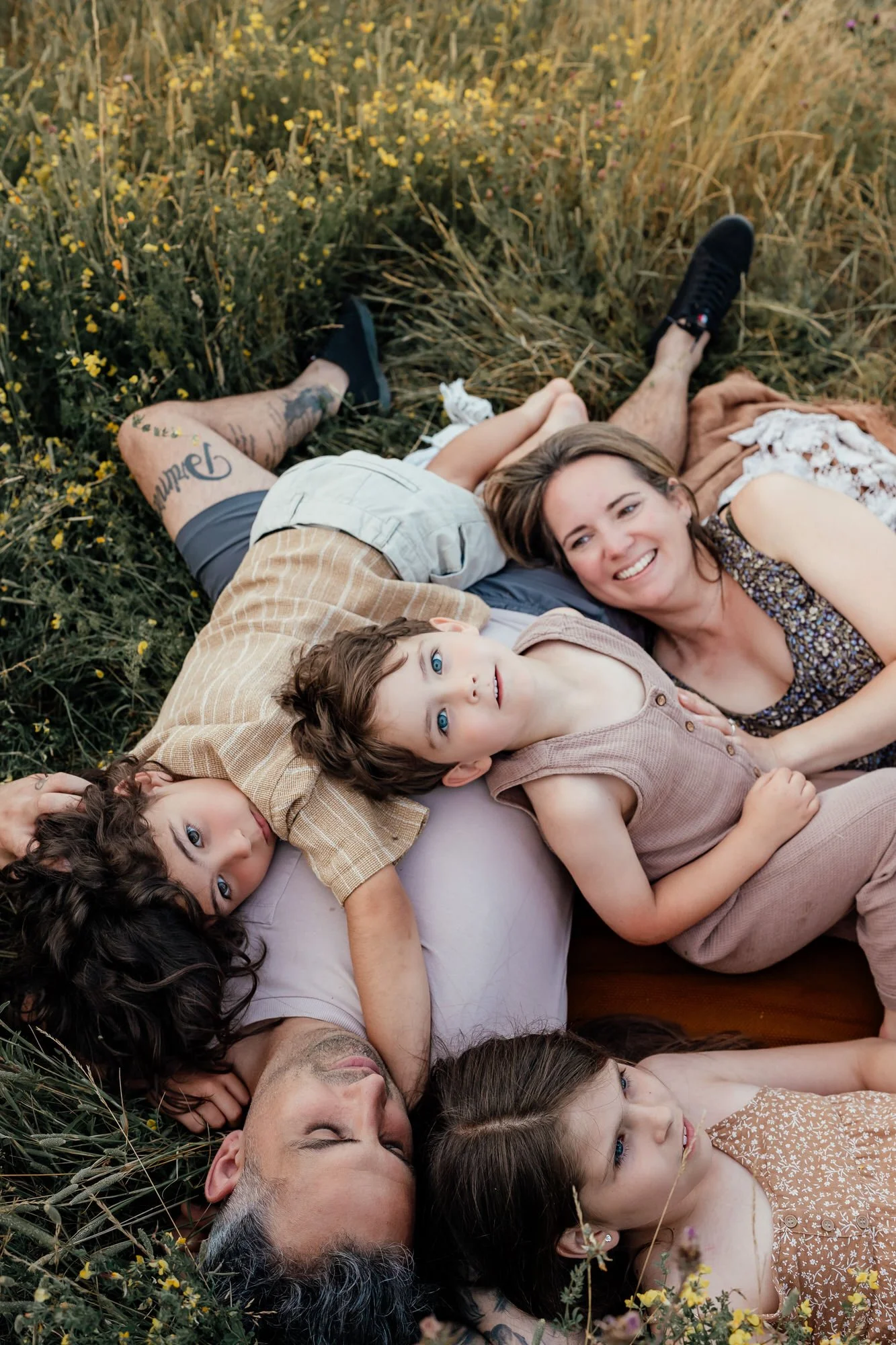 family of two boys, two parents, one girl are all lying on the floor in the grass in summer over the dad's chest looking at the camera