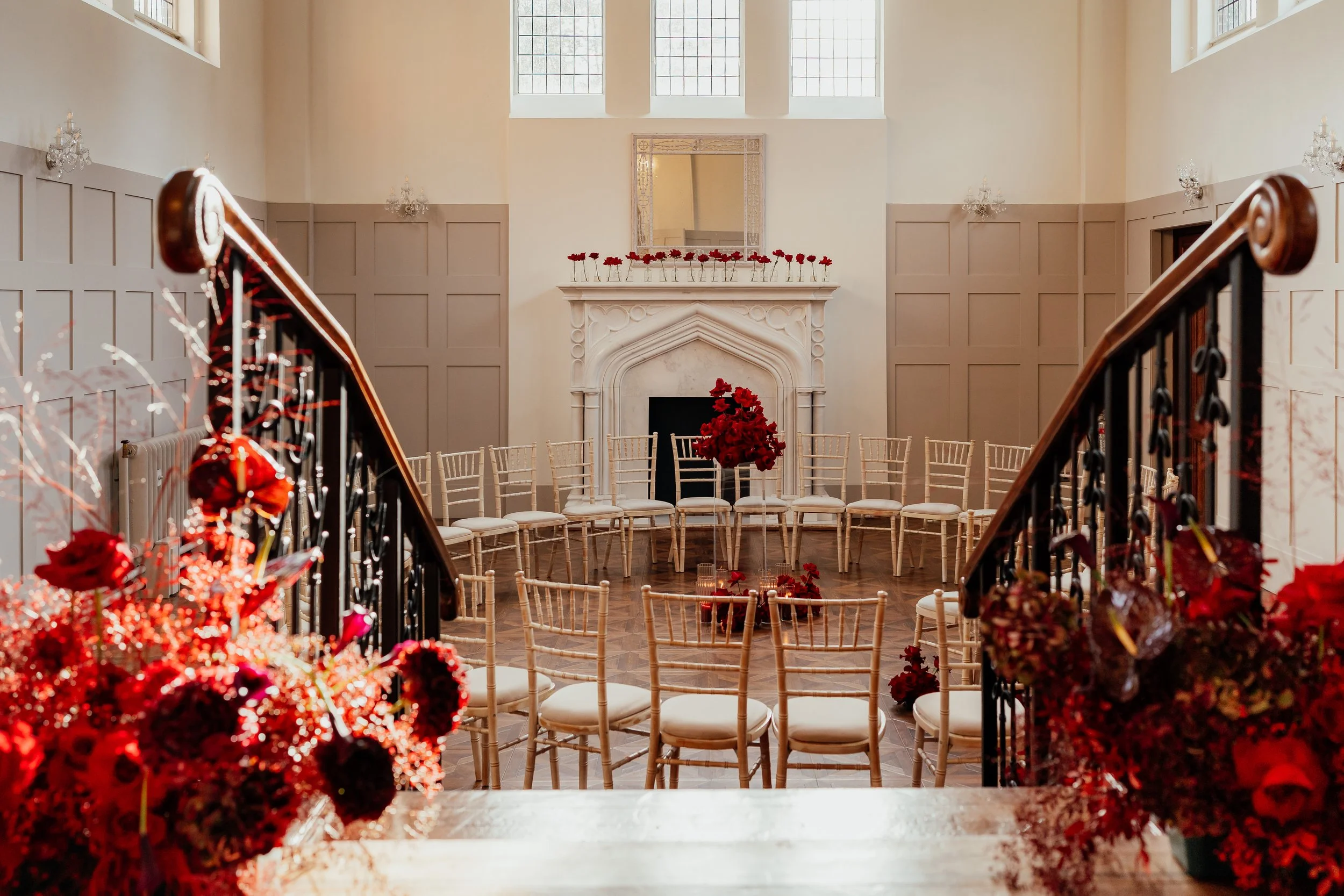 view down to a circular wedding ceremony set up, no people, view through the stairs down to the ceremony room which is panelled in neutral wood panelling with a large marble fireplace on the back wall. Room dressed with dark red flowers throughout