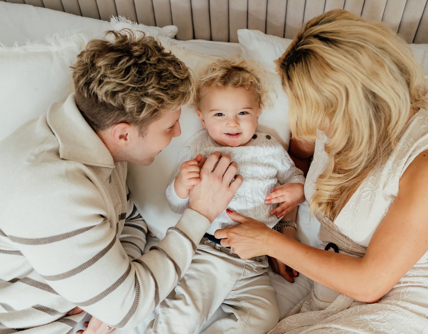 blonde curly haired toddler is central to the image, being tickled by his parents either side of him on a bed, viewing them from above