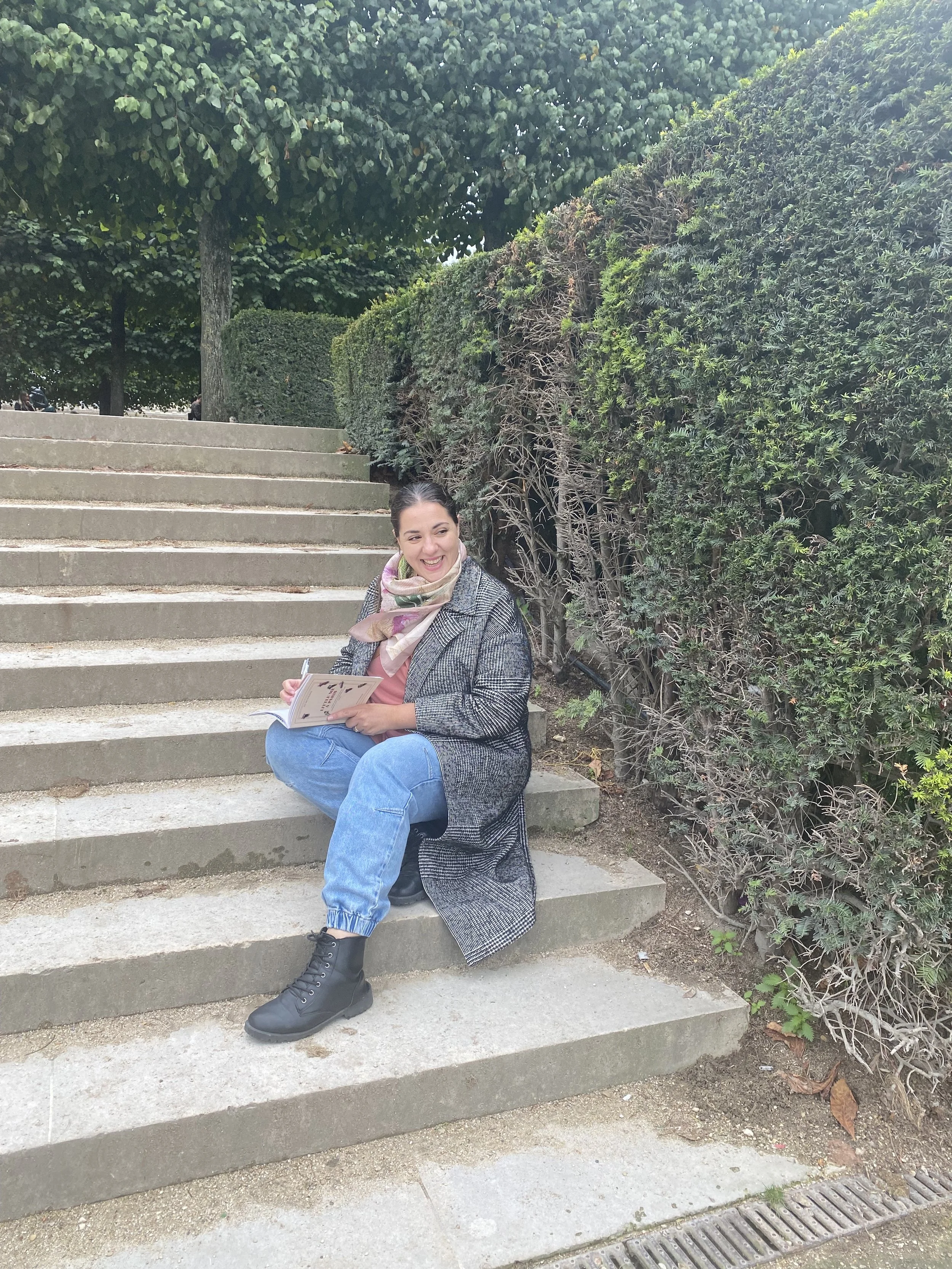 Une femme assise sur des escaliers en pierre, tenant un carnet et souriant dans un parc arboré.