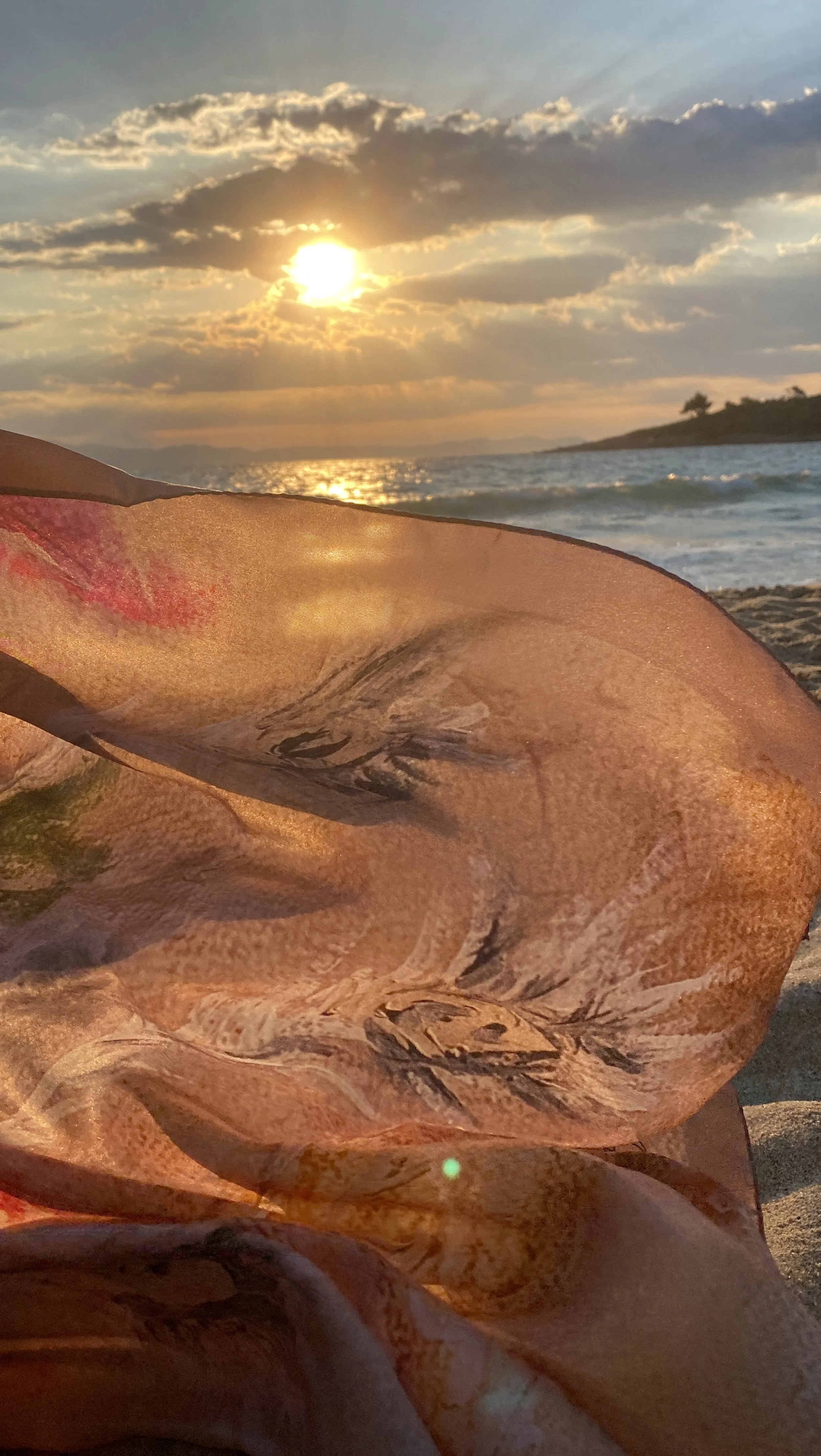 Coucher de soleil sur une plage avec un voile transparent et coloré sur le sable, vue sur la mer et un ciel partiellement nuageux.