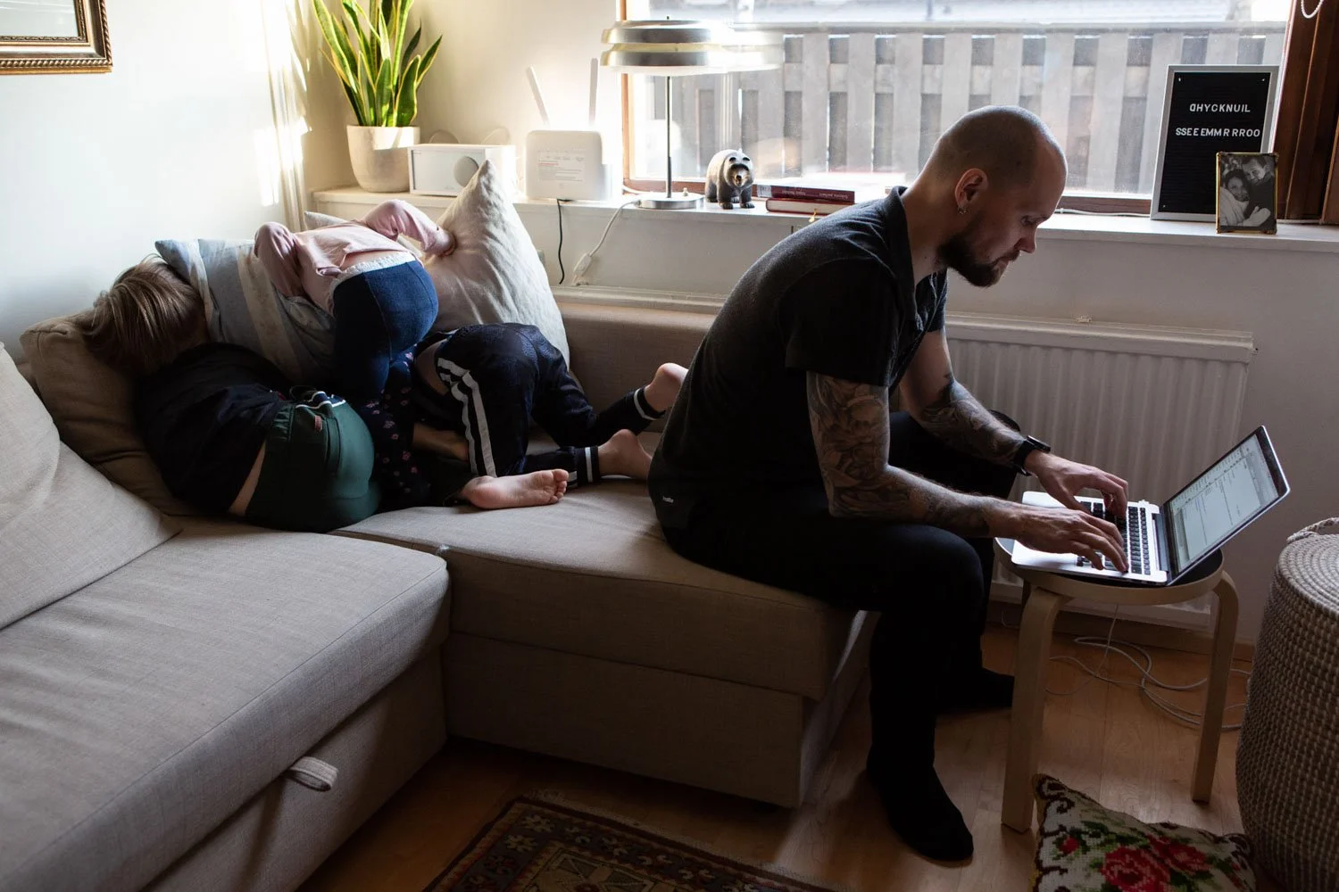 Mies istuu sohvan reunalla ja kirjoittaa tietokoneella. Lapset leikkivät sohvan nurkassa. Dokumentaarinen valokuva. Poikkeustila2020 -projektista.