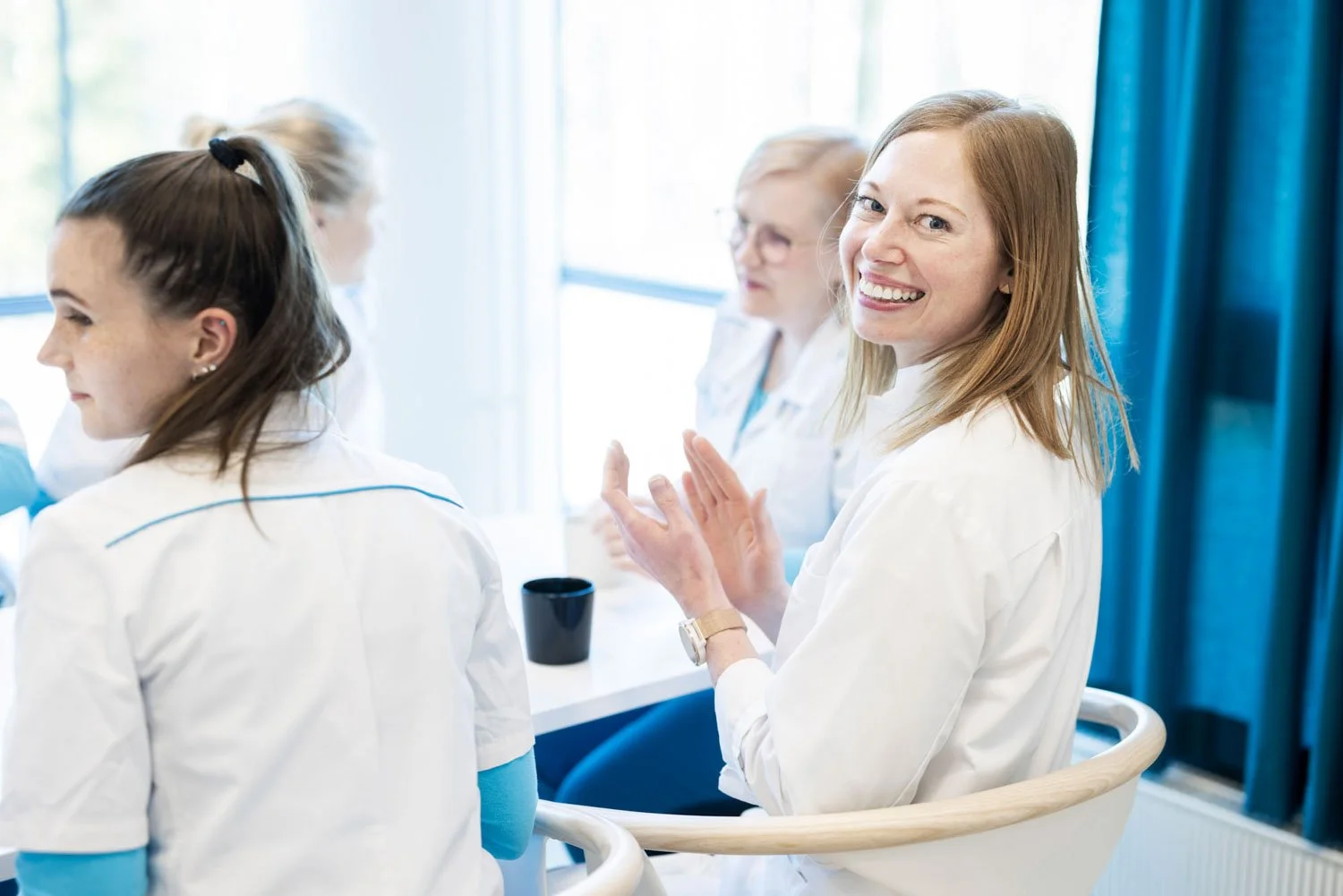 Nainen istuu pöydän ääressä, taputtaa ja katsoo hymyillen kameraan. Ympärillä myös muita ihmisiä.