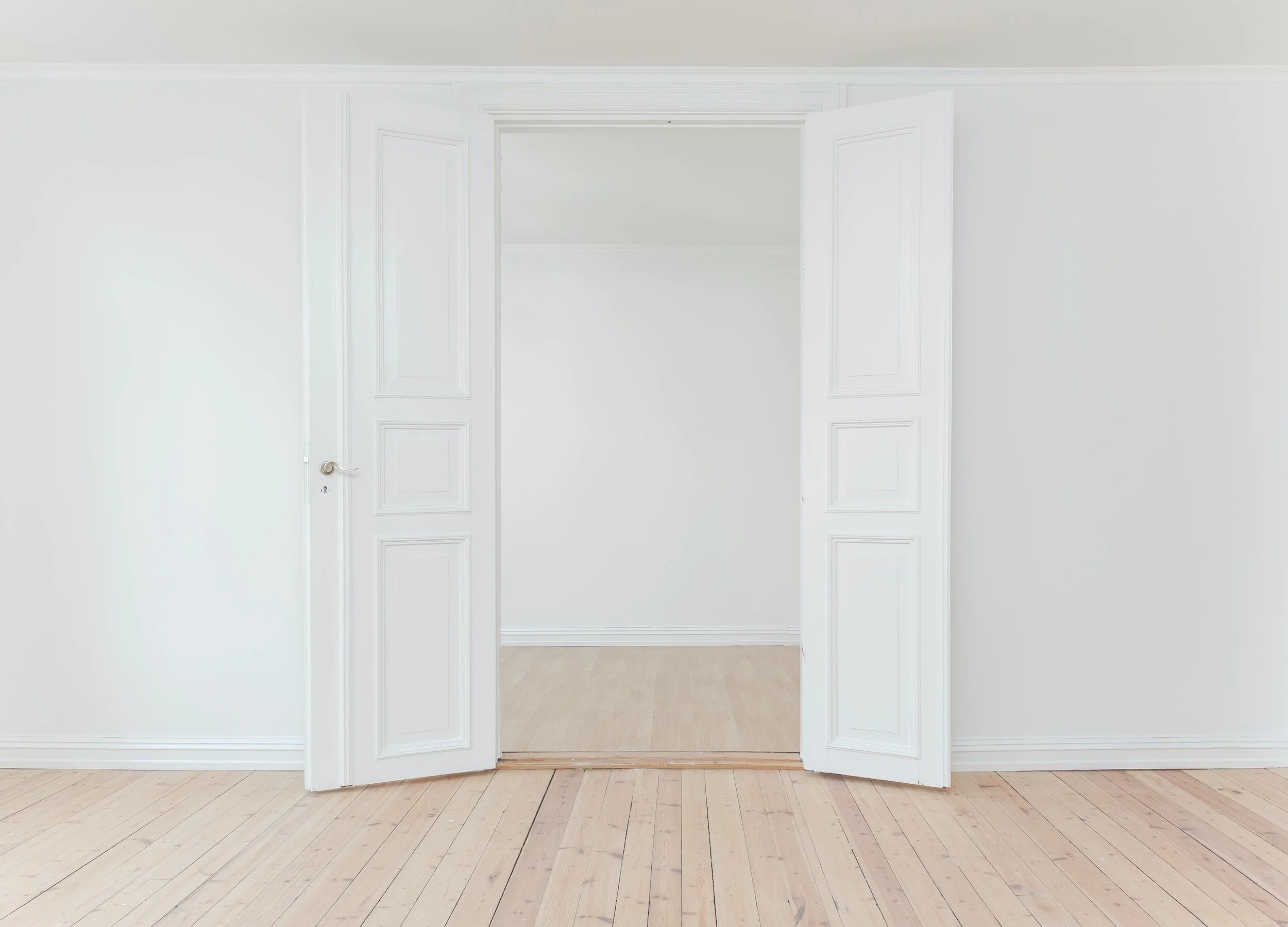 Open white double doors in an empty room with light wooden floor and white walls.