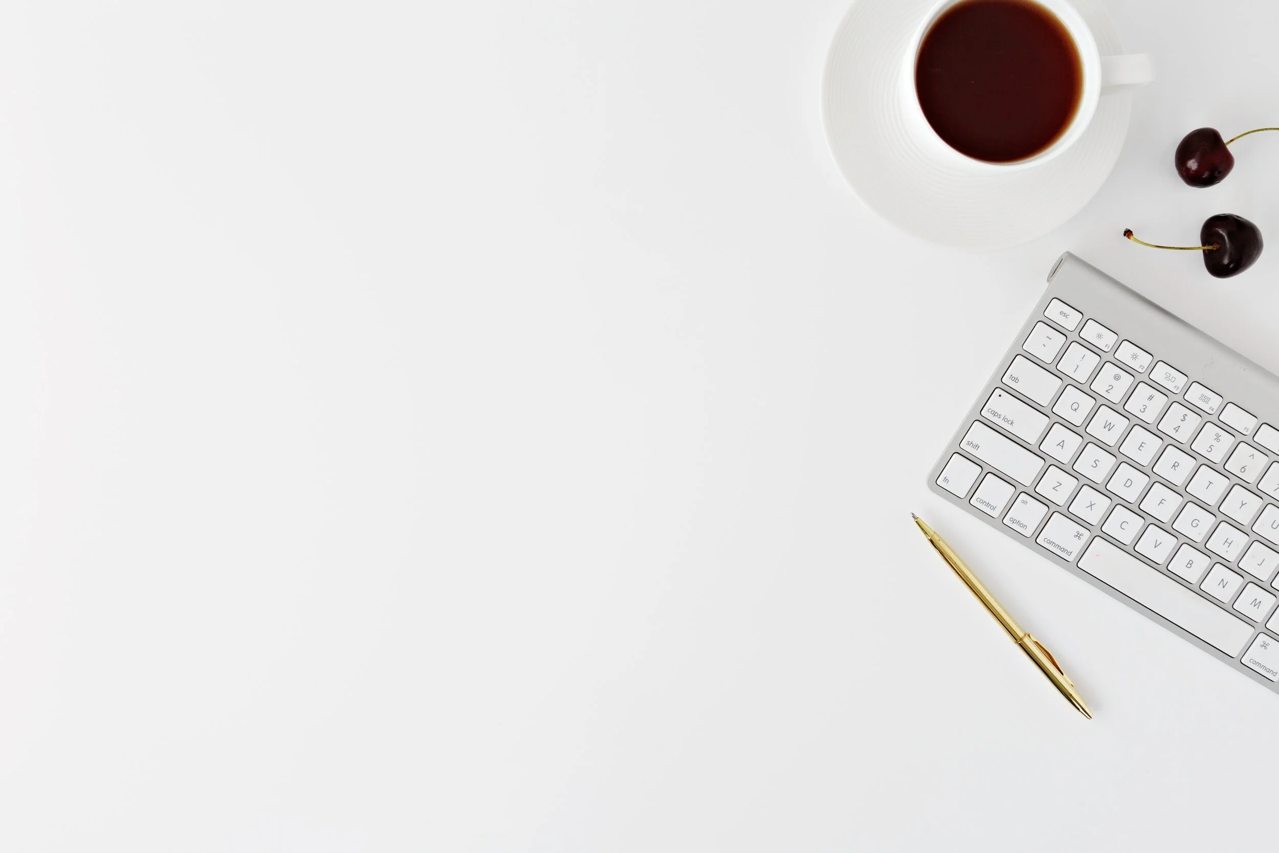 Minimalist workspace with coffee cup, keyboard, pen, and cherries on white background.