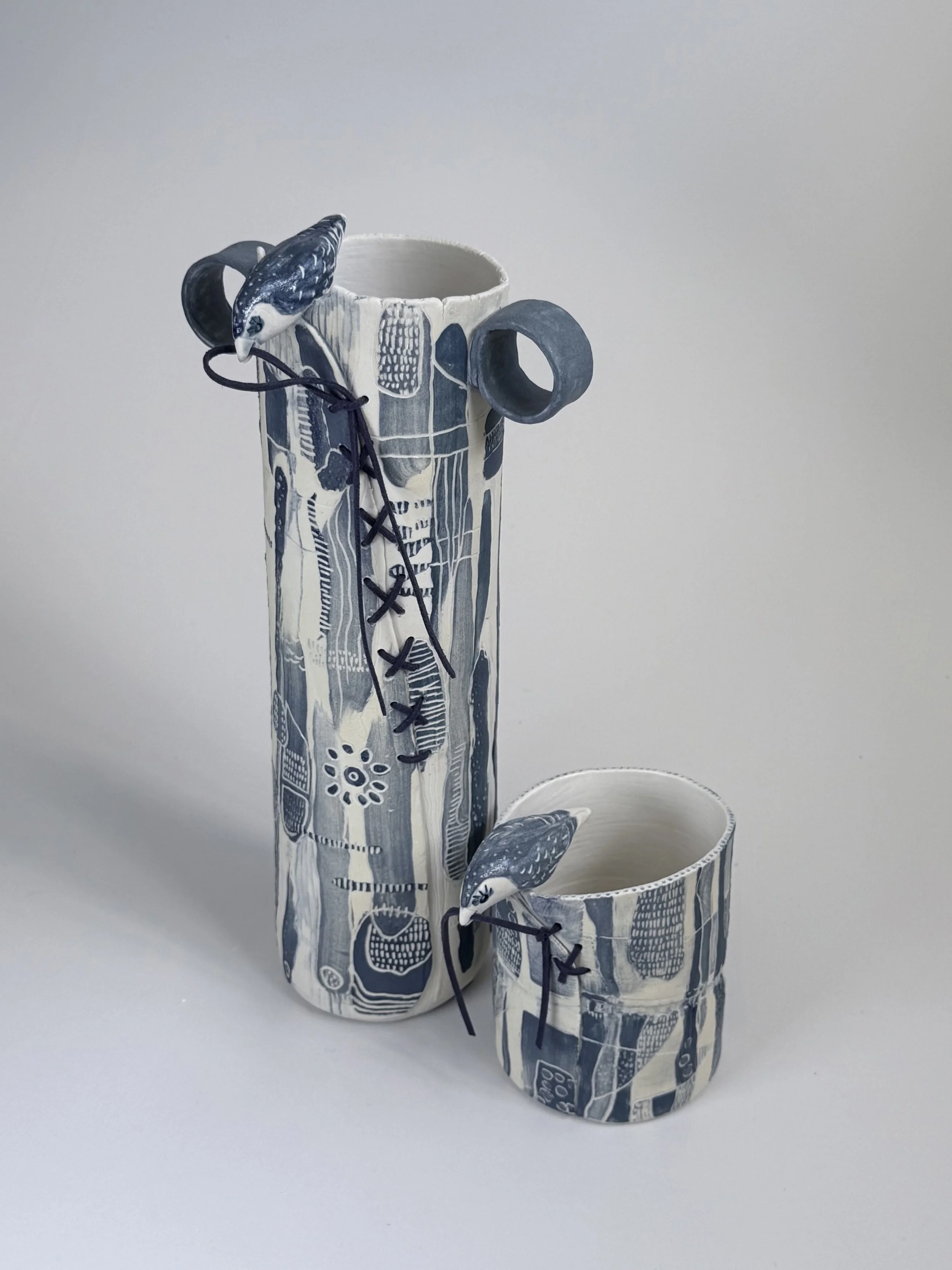 Two ceramic vessels, one tall the other short. Birds sitting on each weaving their vessel closed with leather cord. Titled The Tall and the Short of It.