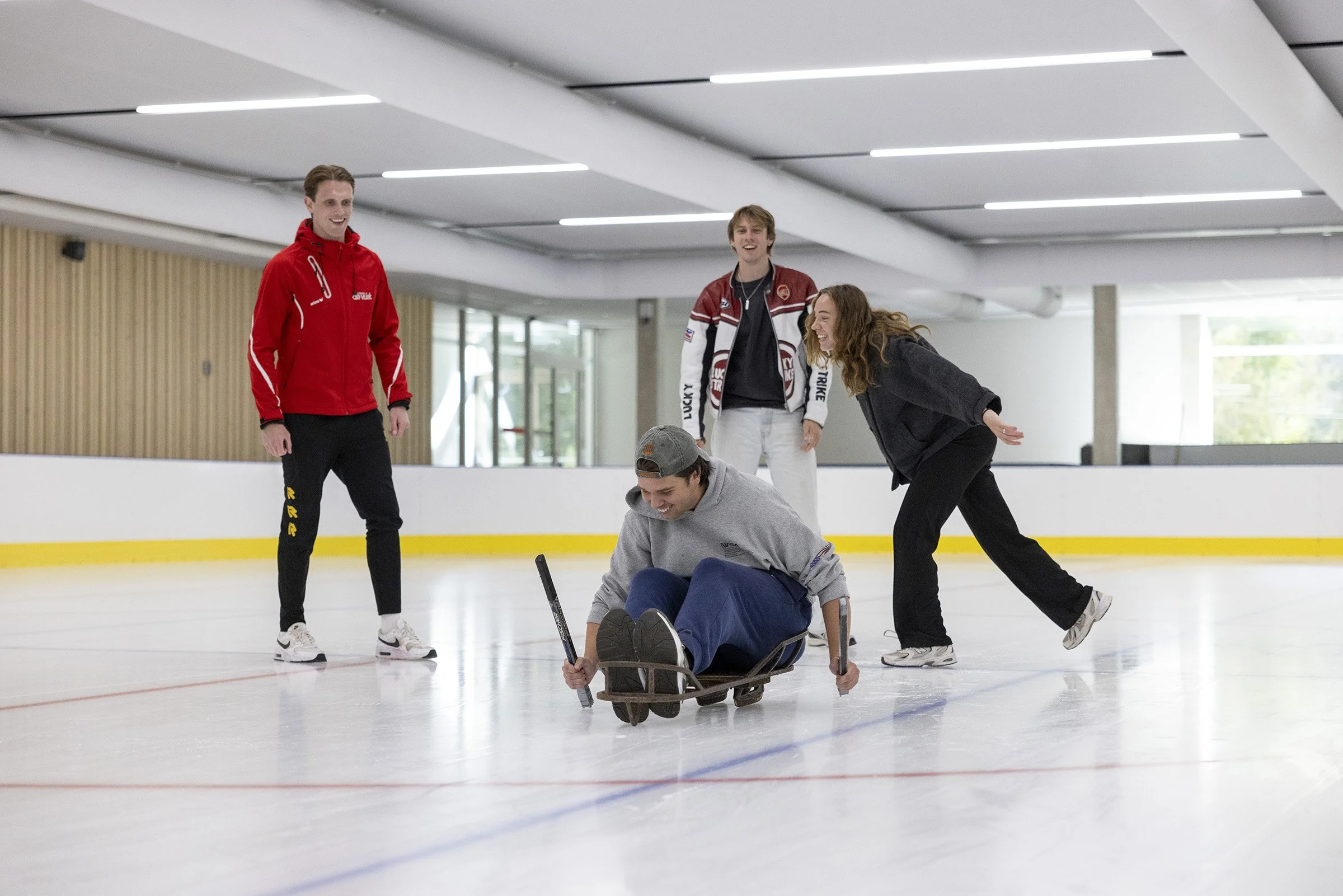 Vijf jongeren op het ijshockeyveld lachen en spelen, één zit in een slee, anderen staan of buigen terwijl ze plezier hebben.