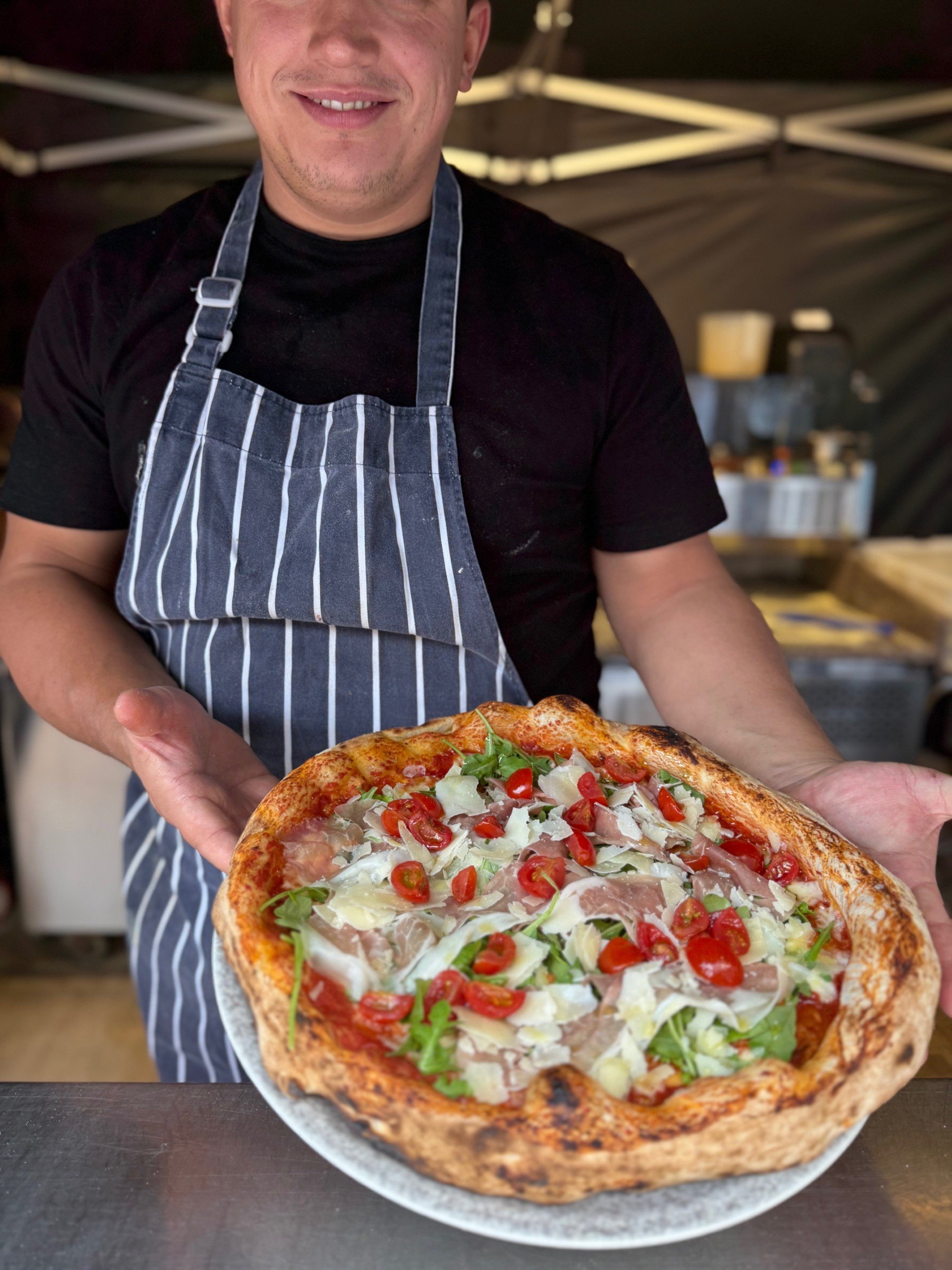 Man holding a freshly baked pizza topped with cherry tomatoes, arugula, shaved cheese, and prosciutto, wearing a striped apron in a outdoor setting.