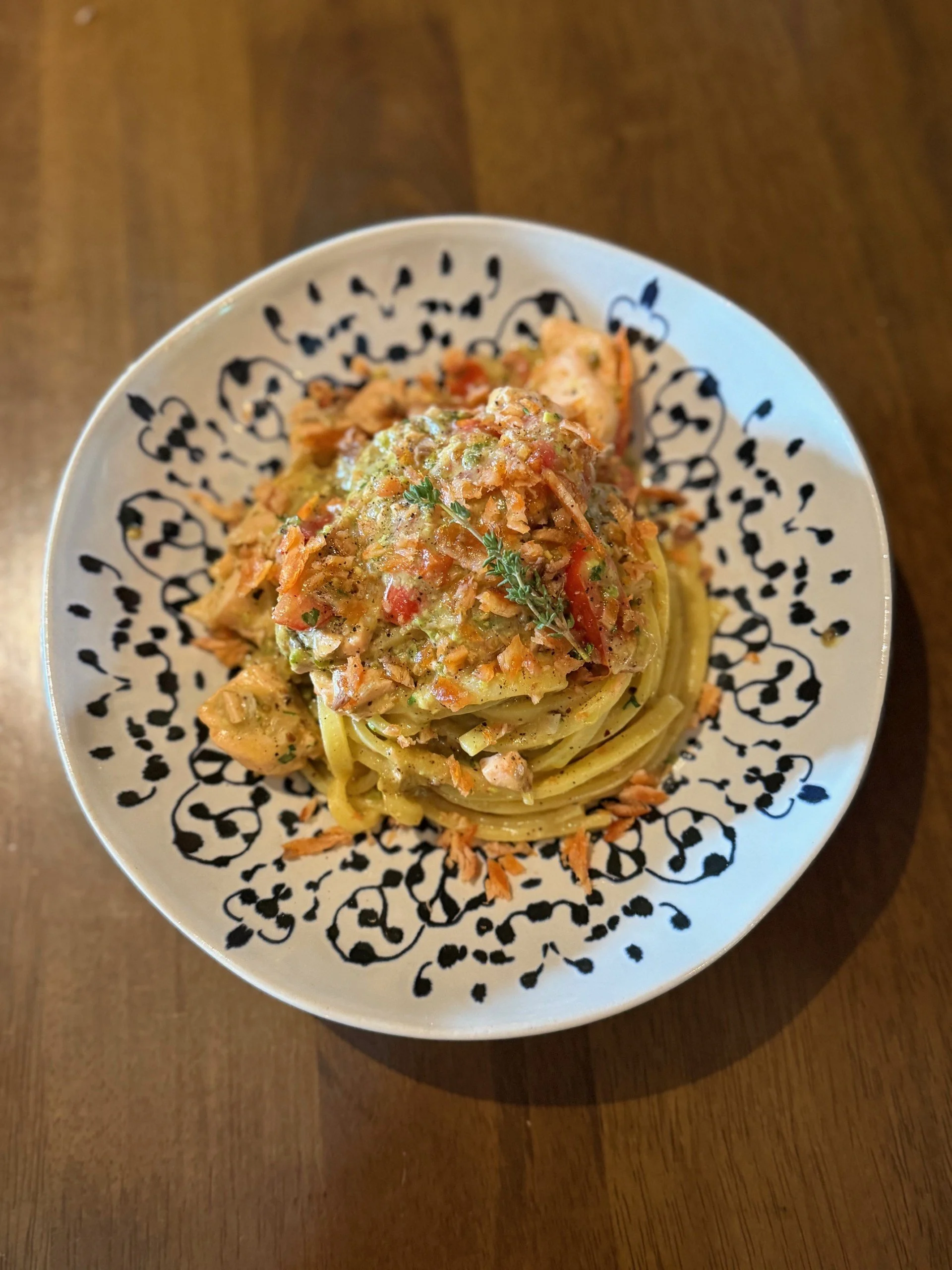 A bowl of creamy salmon and tomato pasta topped with herbs, served on a decorative black-and-white patterned plate on a wooden surface.