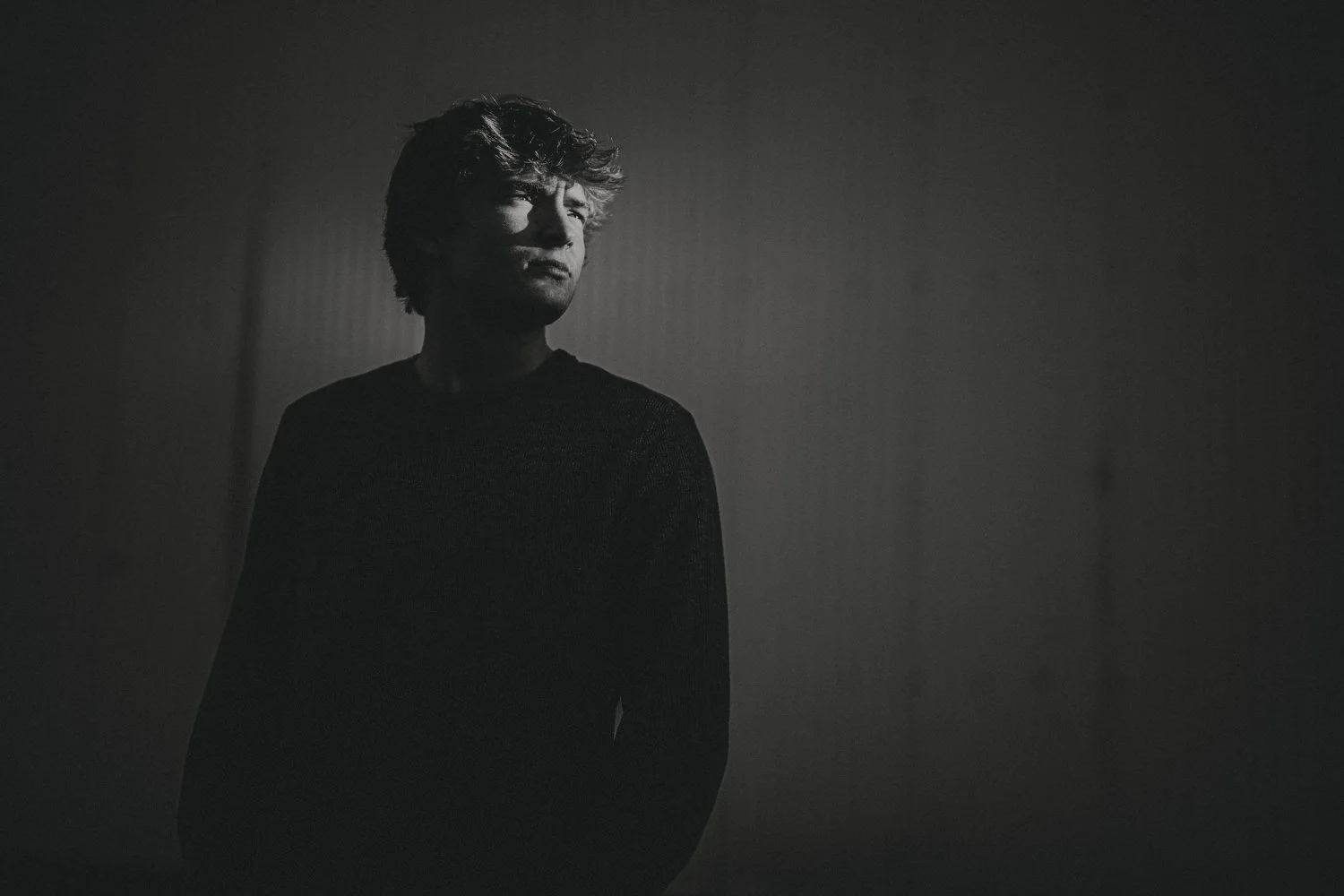 A black and white photo of a young man with wavy hair, standing in front of a plain wall, looking to his left with a serious expression.