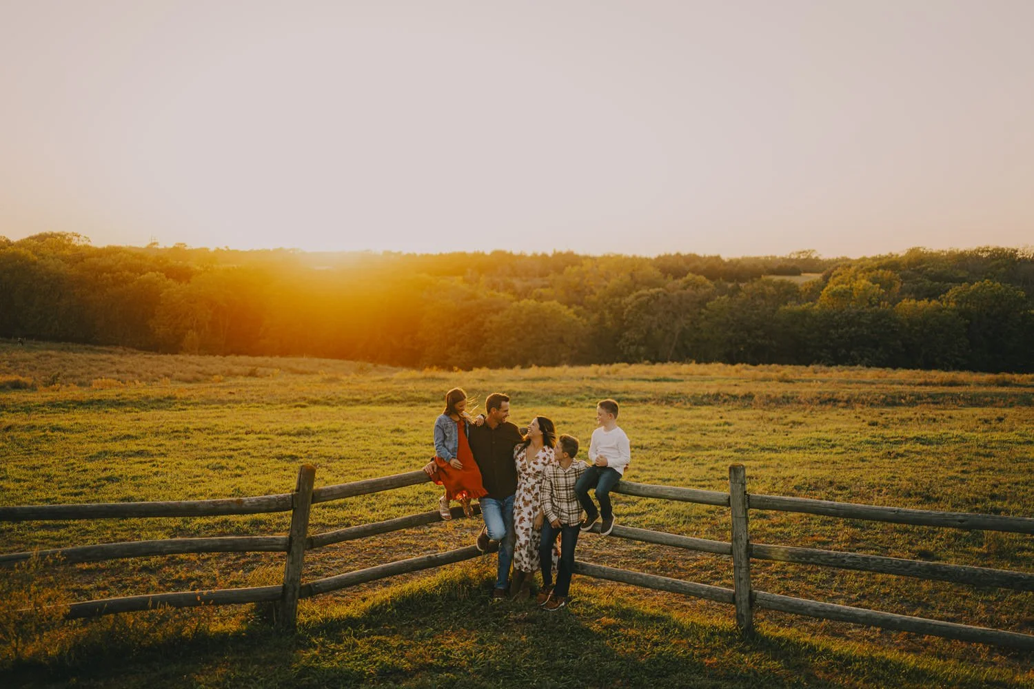 A family of six, including two adults and four children, enjoying sunset in a field by a wooden fence.