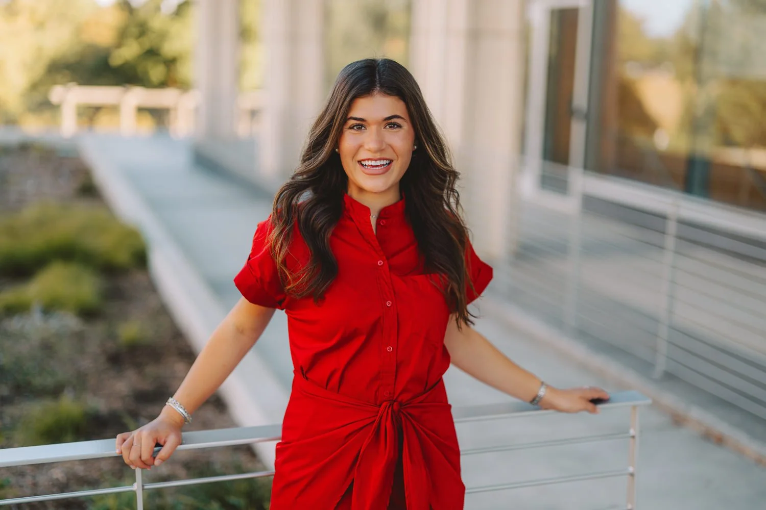 A woman with long dark hair, wearing a red dress, smiling and leaning on a white railing outdoors.