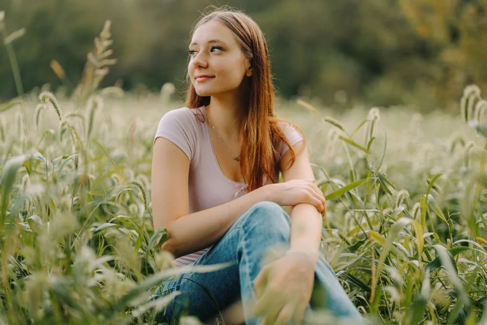 A young woman with long reddish hair sitting in a field of tall grass, looking off to the side with a slight smile.