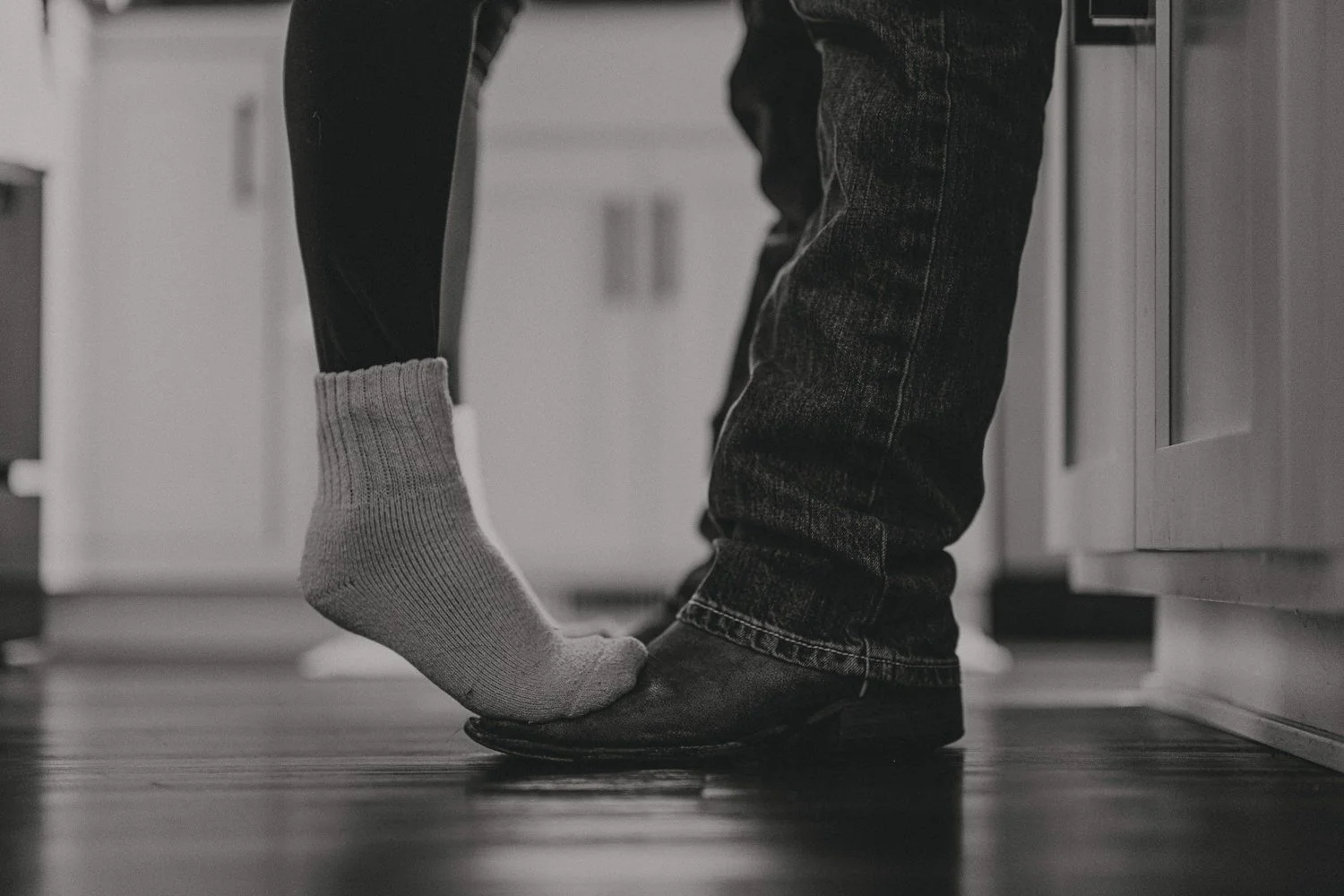 A person wearing dark jeans and black shoes is standing in a kitchen, about to step on a person wearing a sock and pants, with the socked foot raised and touching the other person's shoe.