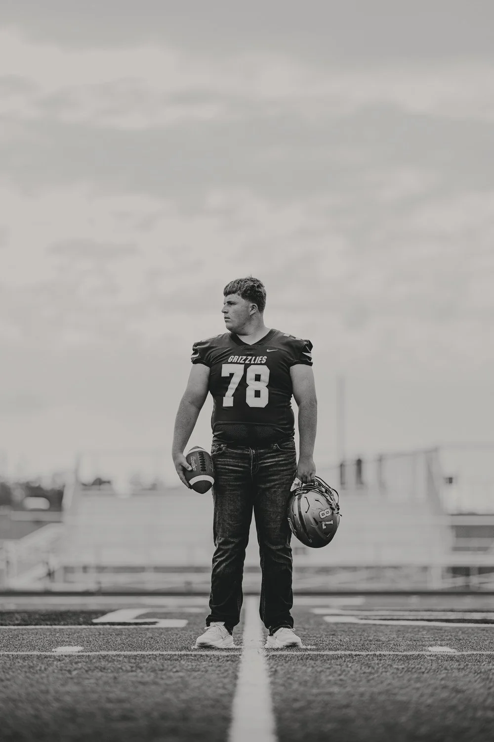 A football player stands on a field, wearing a dark jersey with the number 78 and the word 'Grizzlies,' holding a football in his right hand and a helmet in his left, with a cloudy sky in the background.