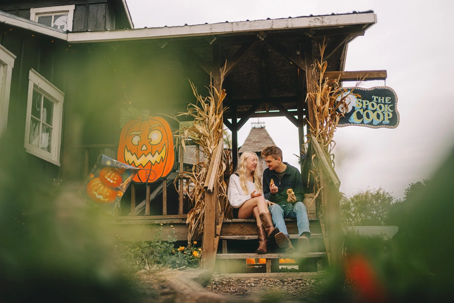 A young couple is sitting on the front porch of a Halloween-themed house, eating cookies and smiling at each other. The house is decorated with orange pumpkin cutouts and dried corn stalks, and there is a sign that reads 'The Spook Coop'.