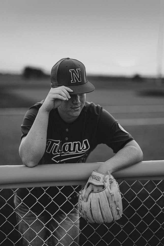 Black and white photo of a young man in a baseball uniform, resting his arm on a fence, wearing a cap and holding a glove, with a baseball field in the background.