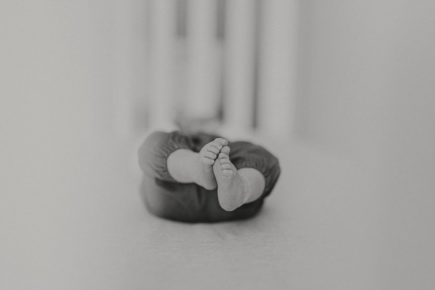 A black-and-white photo of a baby lying on its stomach with its legs bent and feet in the air, on a smooth surface.