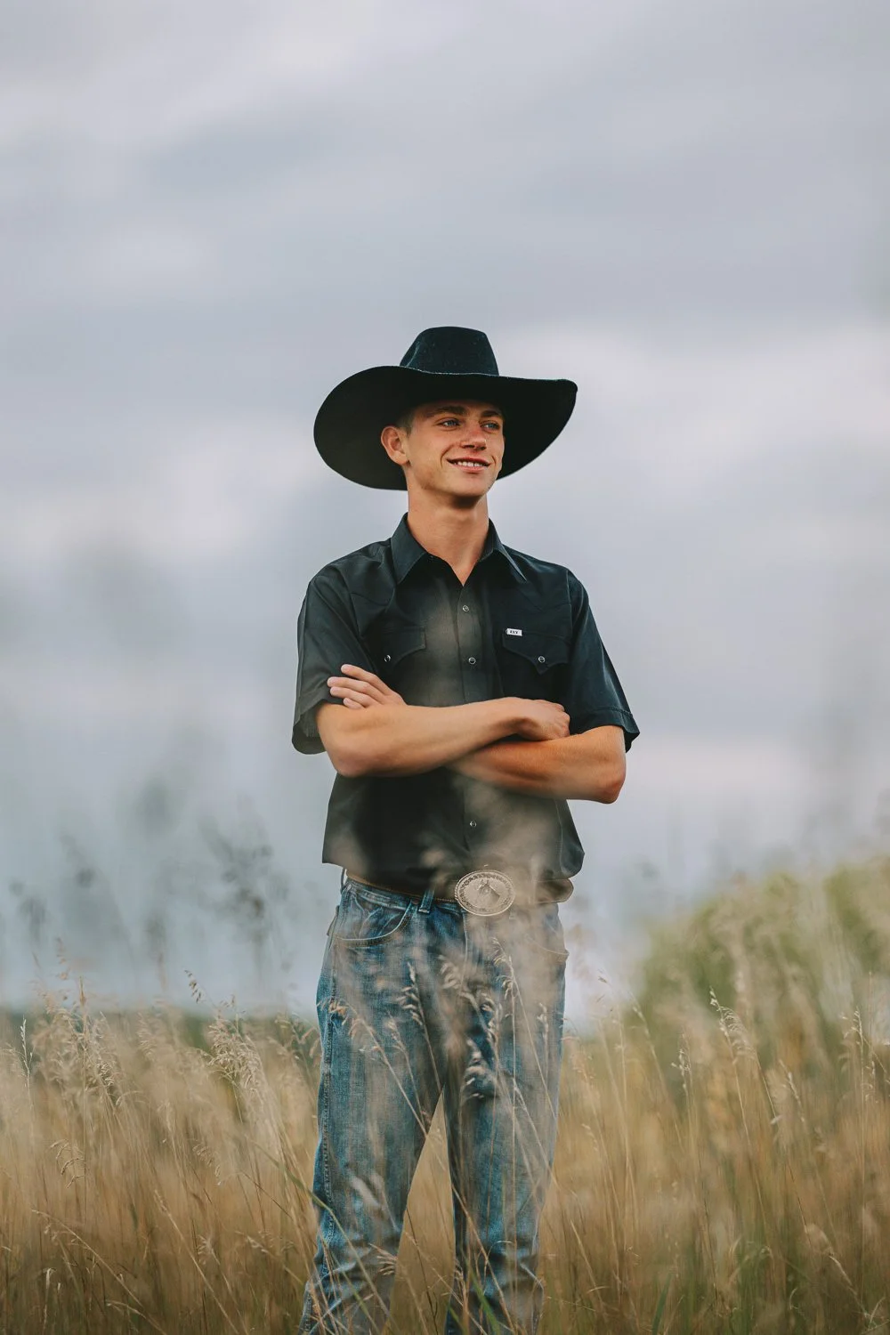 Young man standing in a field of tall grass, wearing a black cowboy hat, black shirt, and jeans, with arms crossed and smiling against a cloudy sky background.
