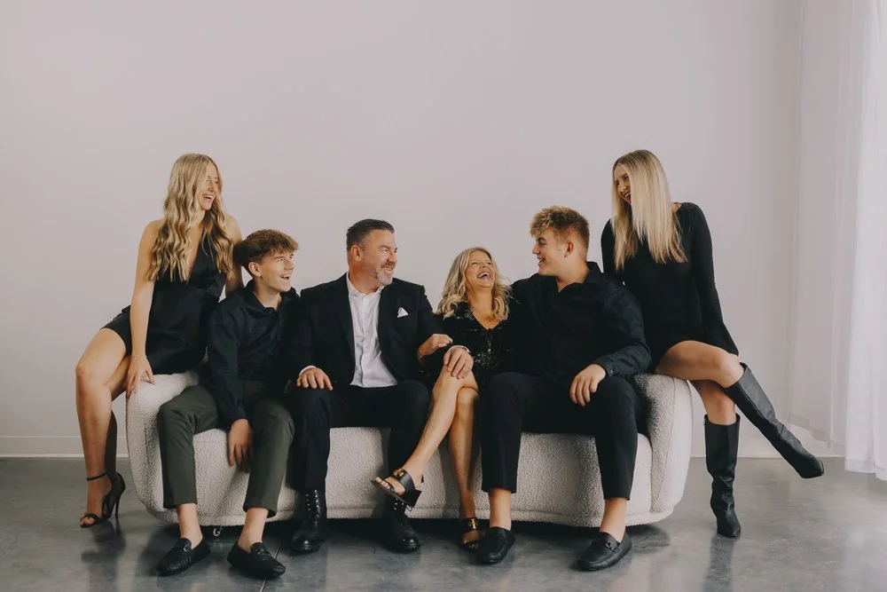 Family of six sitting and standing on a sofa, smiling and sharing a joyful moment in a bright room.