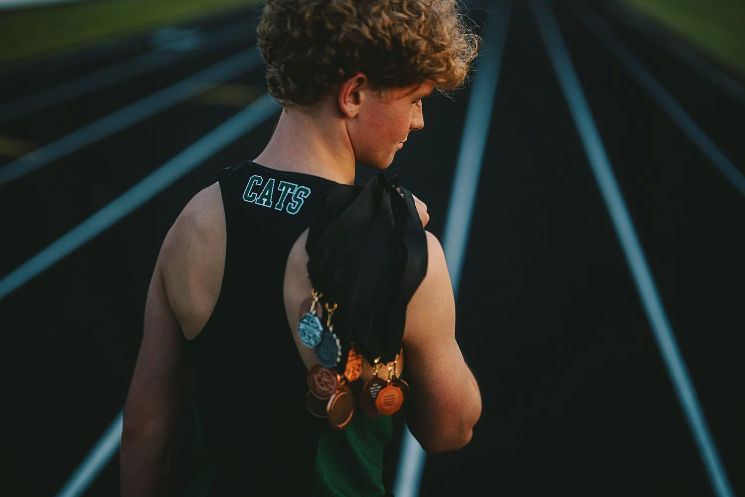A young person with curly hair wearing a sports jersey with 'CATS' on the back, holding a black jacket with medals hanging from their shoulder, standing on a running track at dusk.