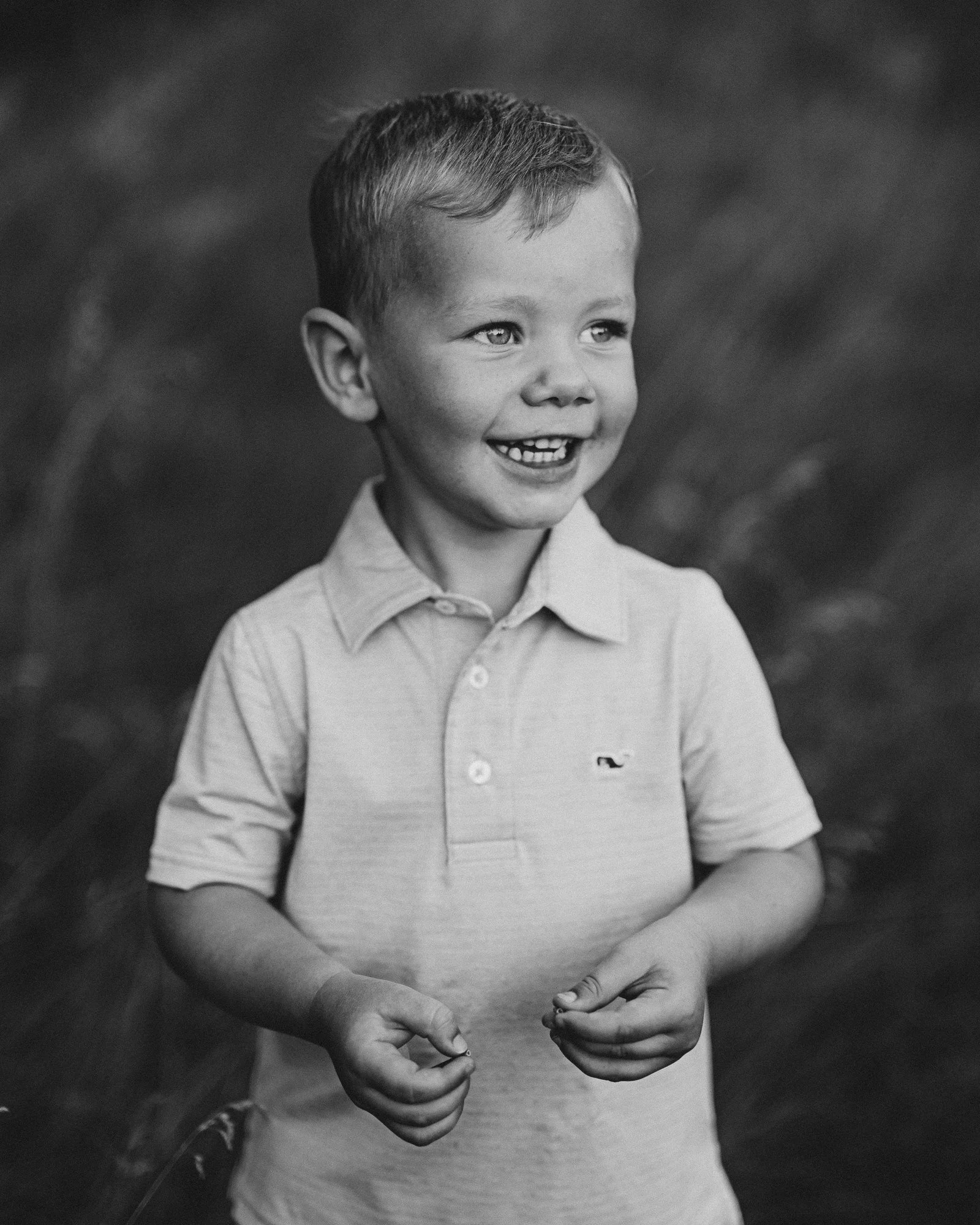 A young boy smiling and laughing outdoors, wearing a collared polo shirt, with a blurred natural background.