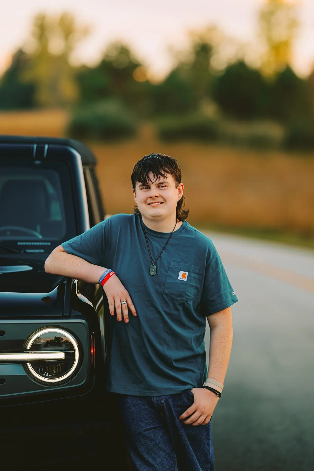 A young man standing outdoors next to a black vehicle, smiling at the camera, with a blurred natural background of trees and sunset.