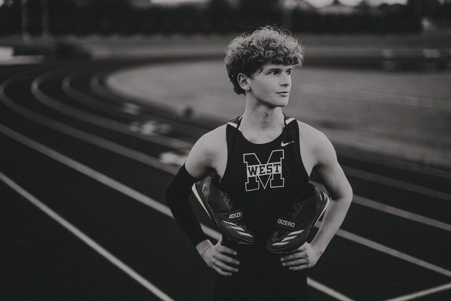 A young male athlete with curly hair standing on a track, wearing a sports shirt with a large 'M' and the word 'WEST' on the front, holding a pair of running shoes.