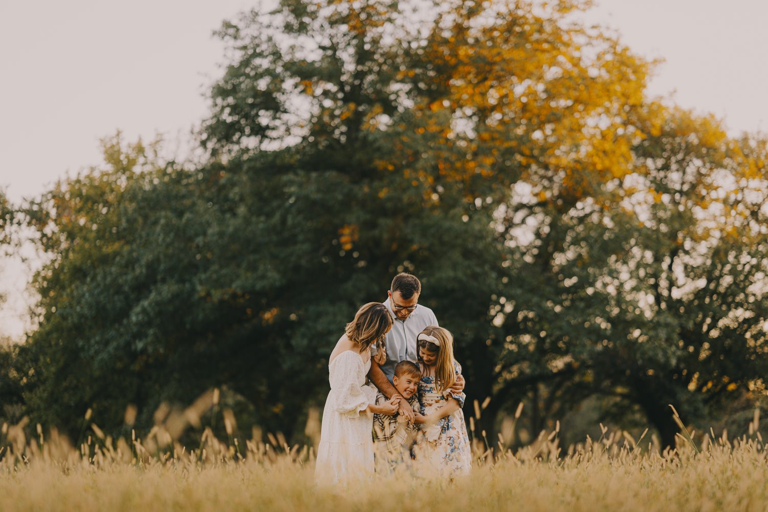 Family of four enjoying a moment together outdoors in a grassy field with large trees and autumn leaves in the background.
