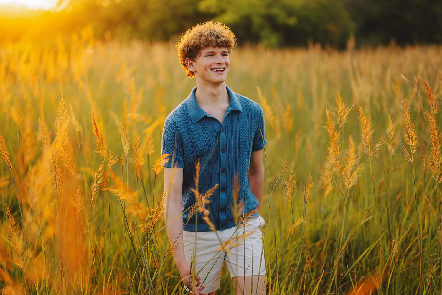 A young man with curly hair wearing a blue shirt and white shorts smiling while walking through a field of tall grass during sunset.