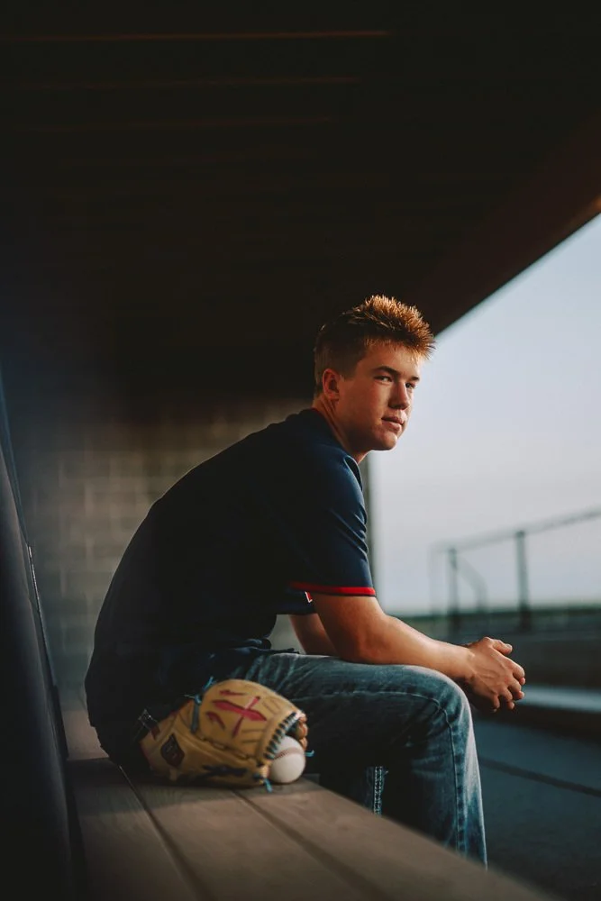 A teenage boy sits on a bench under a bridge, wearing a dark shirt with red accents, jeans, and holding a baseball glove. The background shows a blurry railing and sky.
