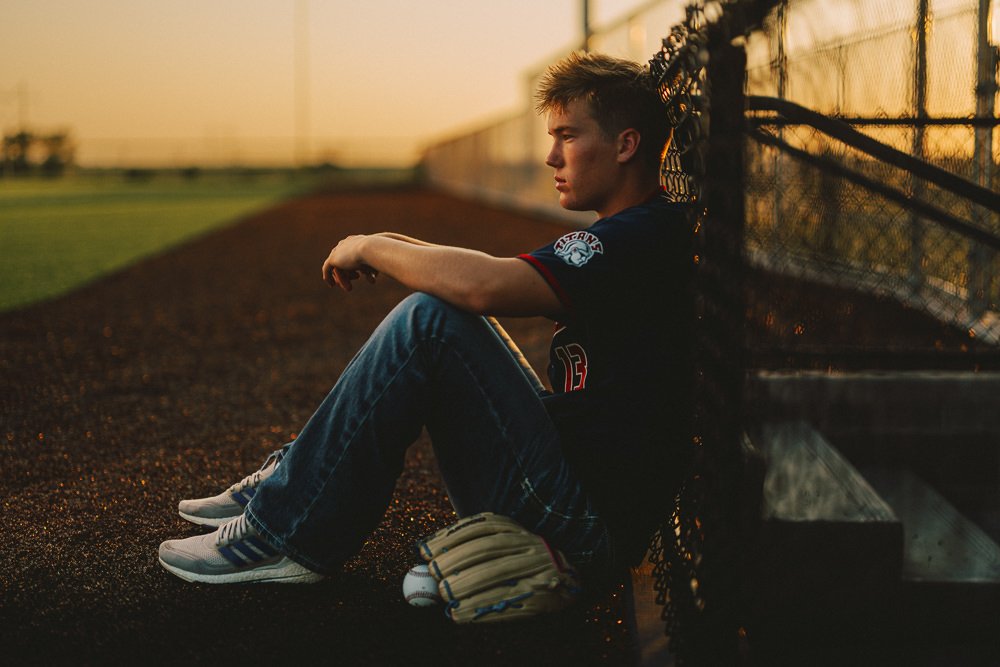 Young male baseball player sitting on the ground, resting against a chain-link fence at sunset, wearing a black jersey, jeans, and sneakers, with a baseball glove on his lap.