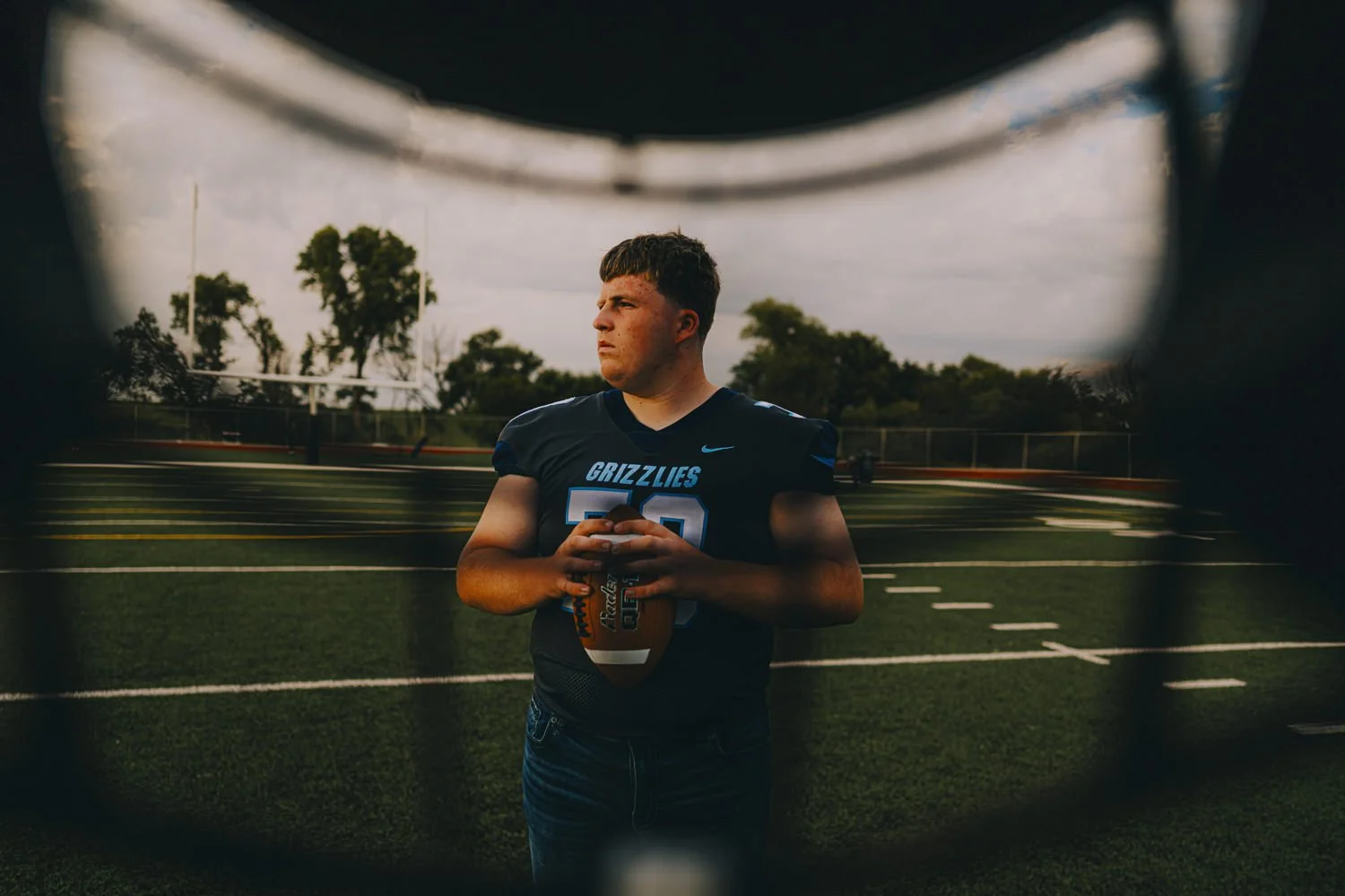 A teenage football player in a black jersey with blue accents, holding a football, standing on a football field seen through a metal fence. Trees and a cloudy sky are in the background.