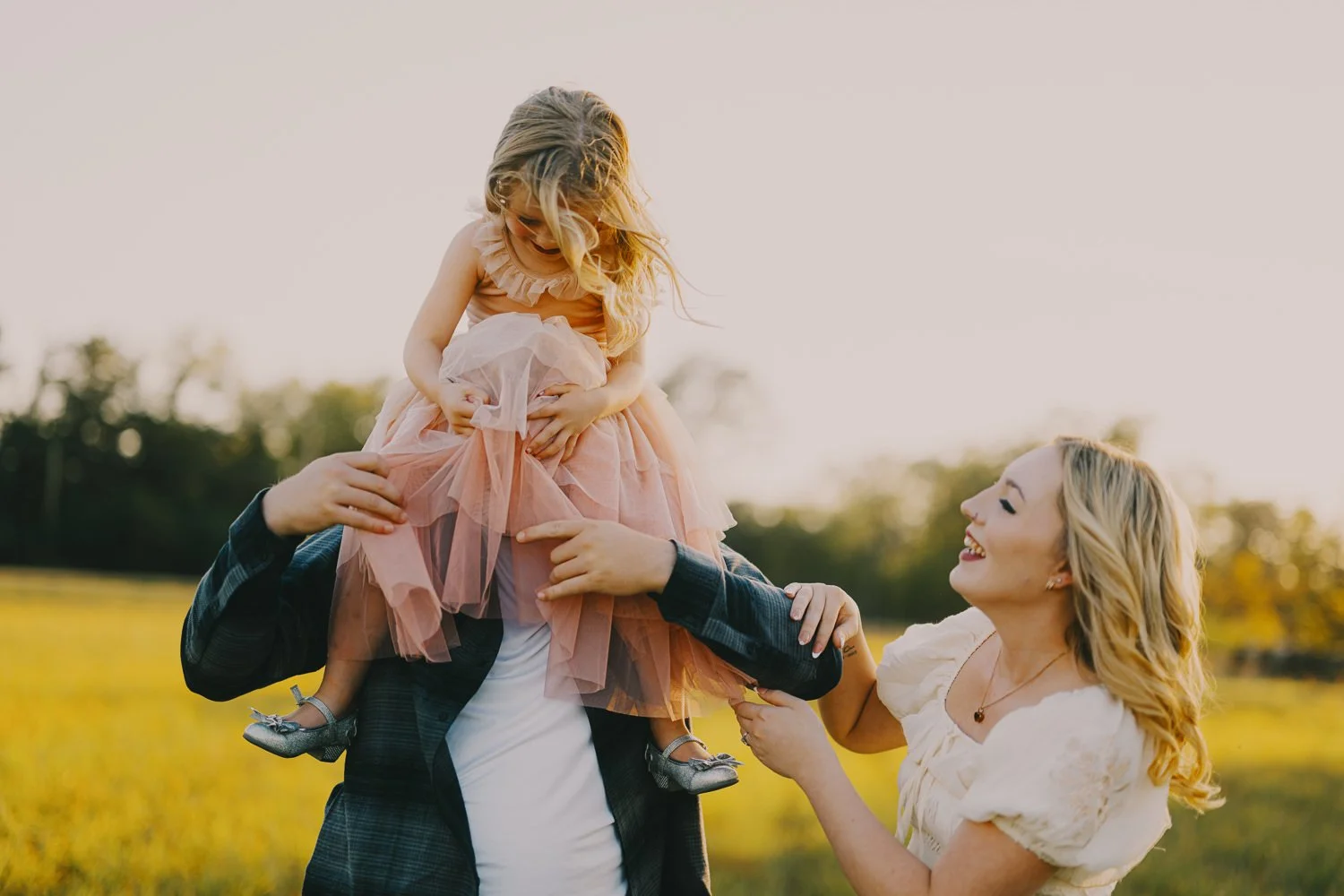 A young girl wearing a pink dress and silver shoes sitting on a man's shoulders, with a woman smiling at her outdoors in a field during sunset.